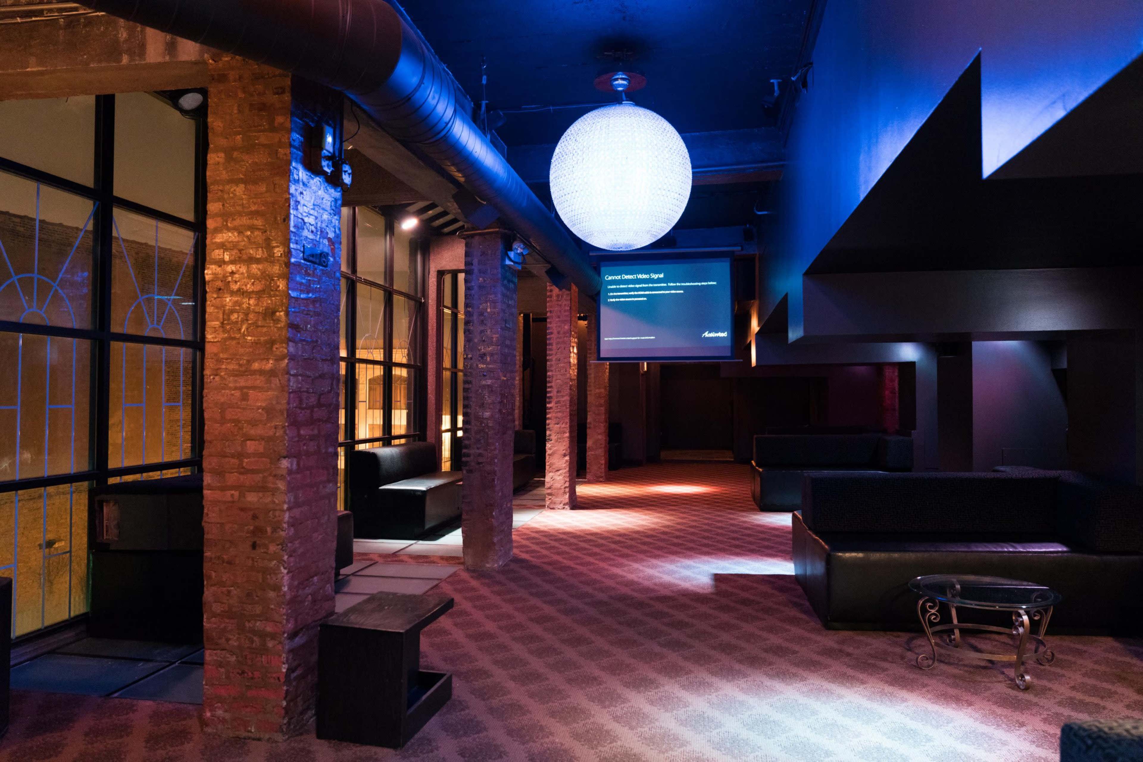 The image shows an empty lounge area featuring exposed brick walls, dark seating arrangements, and a large spherical light fixture hanging from the ceiling.
