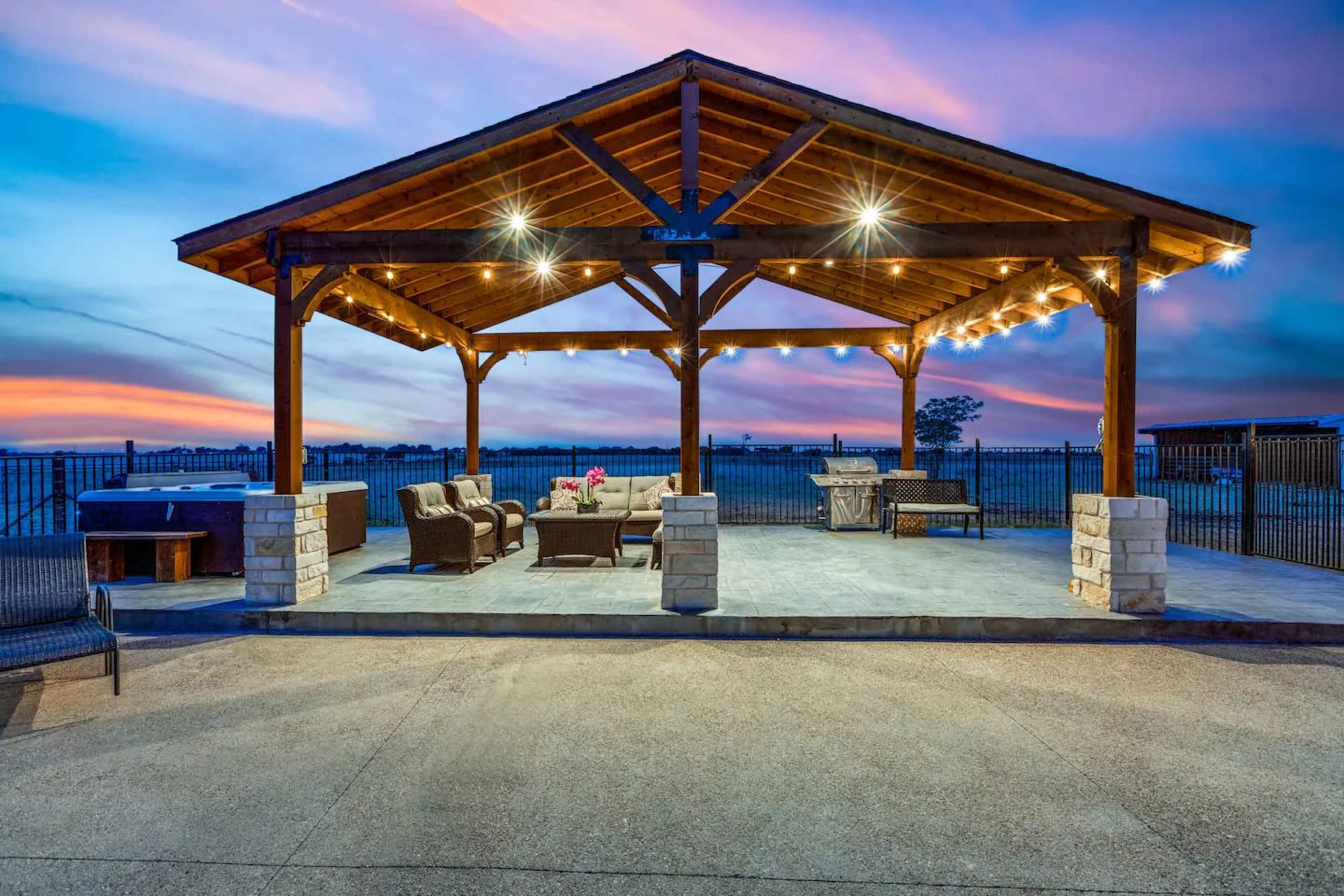 A wooden gazebo with string lights, furnished with outdoor seating, overlooking a scenic sunset.