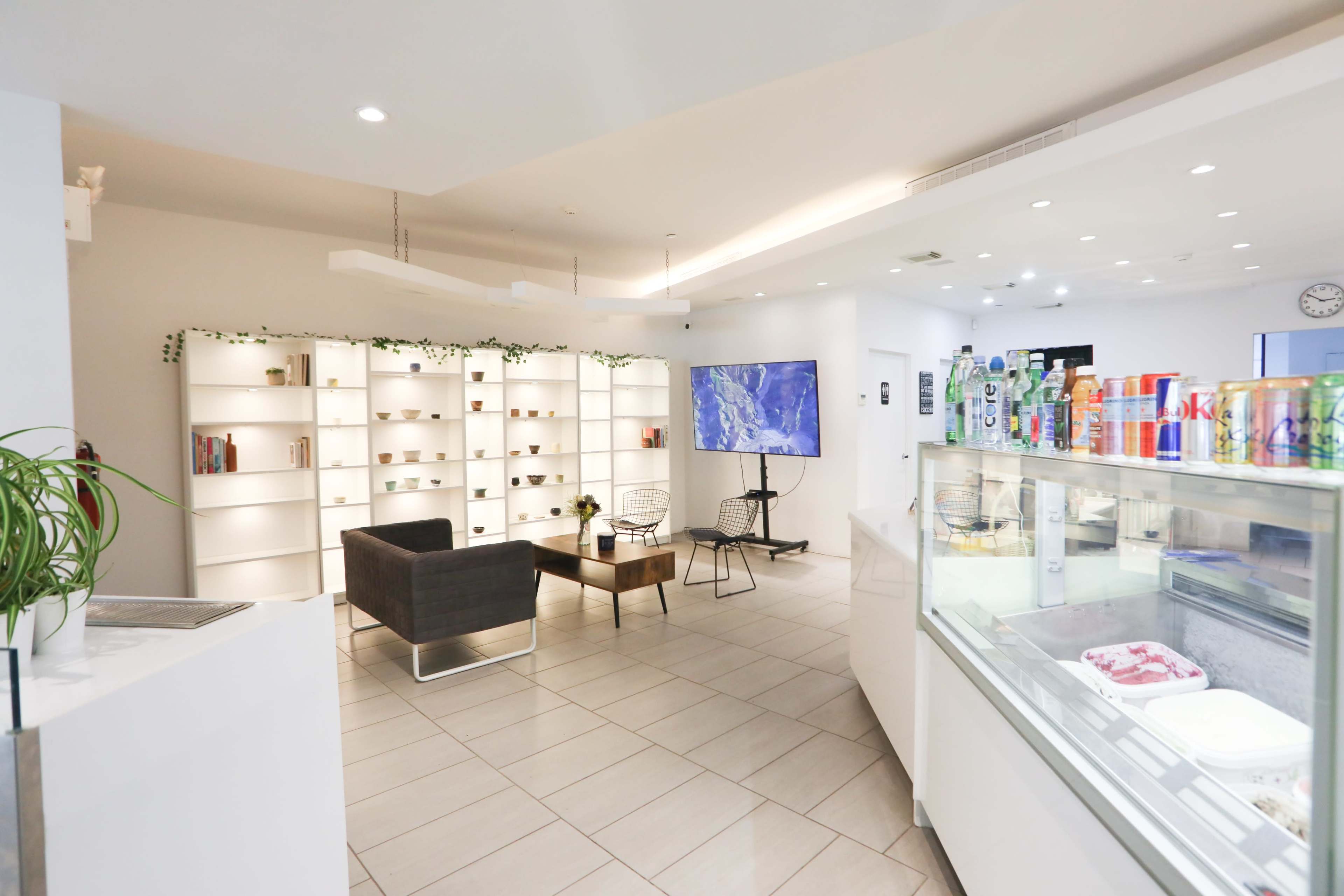 A modern café interior with a seating area, a display case for desserts, a large screen, and shelves showcasing various items.