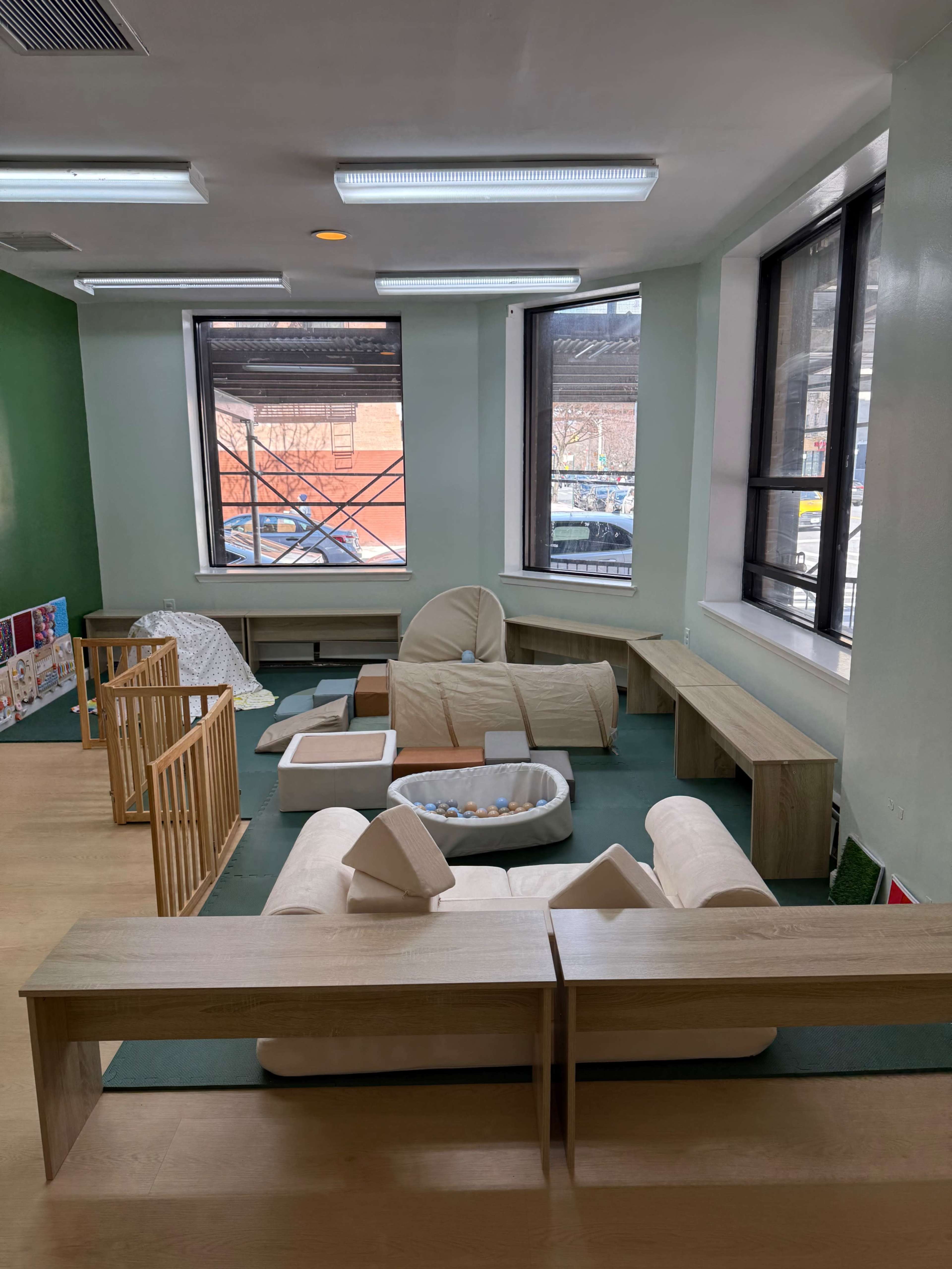 The image shows a playroom with soft furniture, play areas, and large windows allowing natural light to enter.