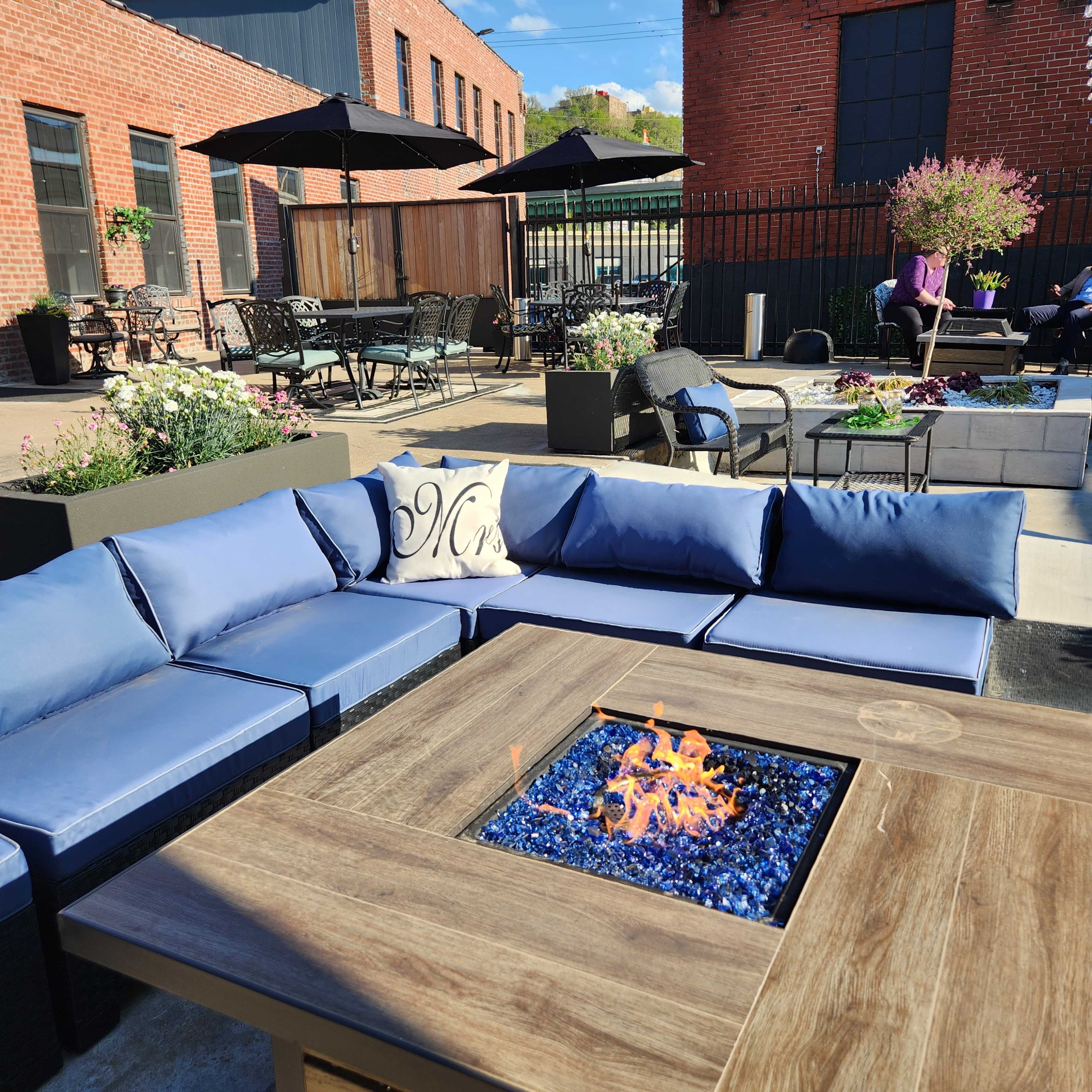 A terrace with a fire pit surrounded by blue cushioned seating, tables, and patio umbrellas near brick buildings.
