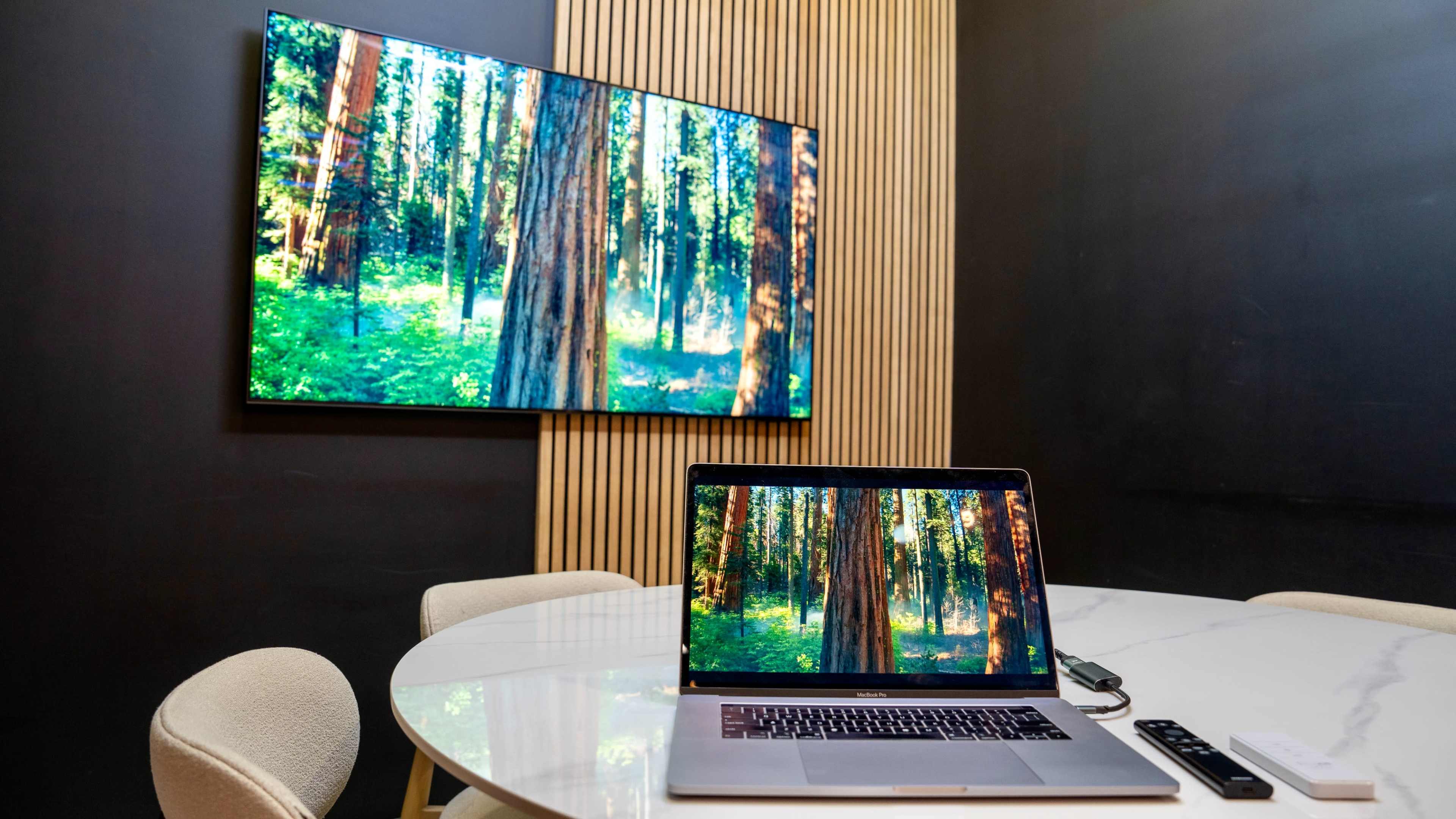 A laptop displaying a forest scene is placed on a table next to a larger television showing the same image in a room with wooden paneling.