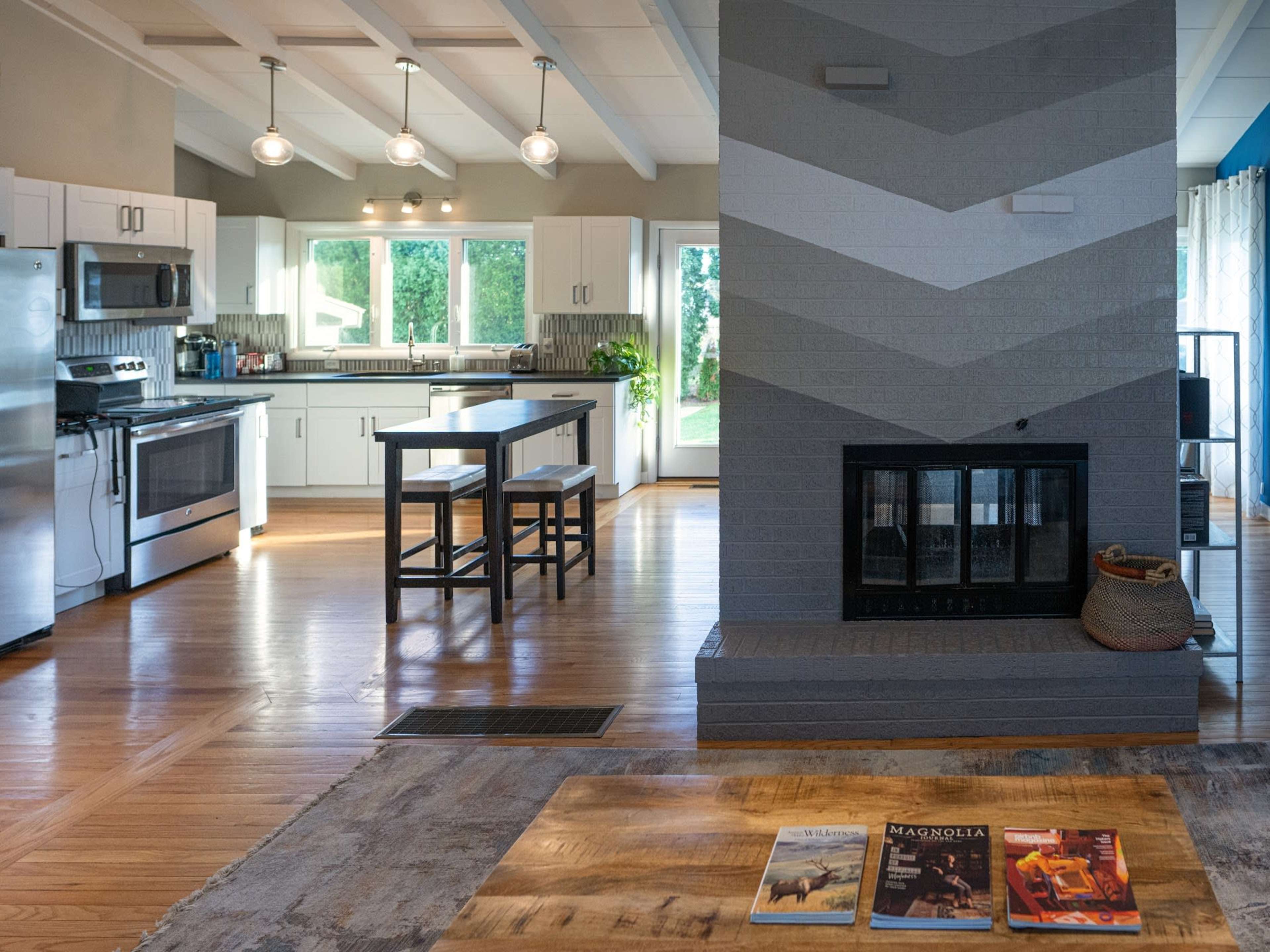 A modern kitchen with stainless steel appliances and a geometric-patterned fireplace, featuring a wooden dining table and magazines on the coffee table.