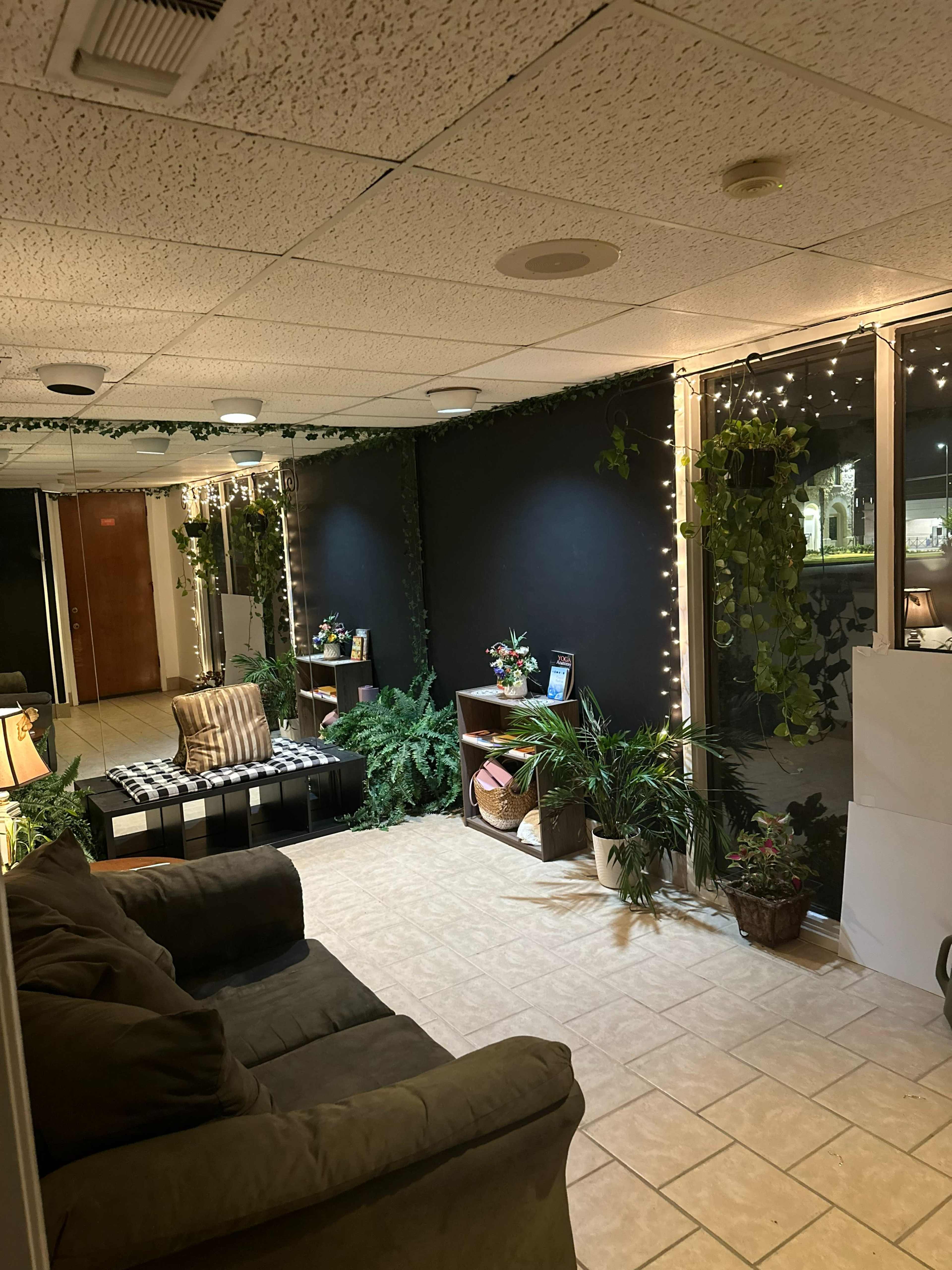 The image shows a cozy indoor space with a sofa and decorative plants, accented by string lights and a dark wall.