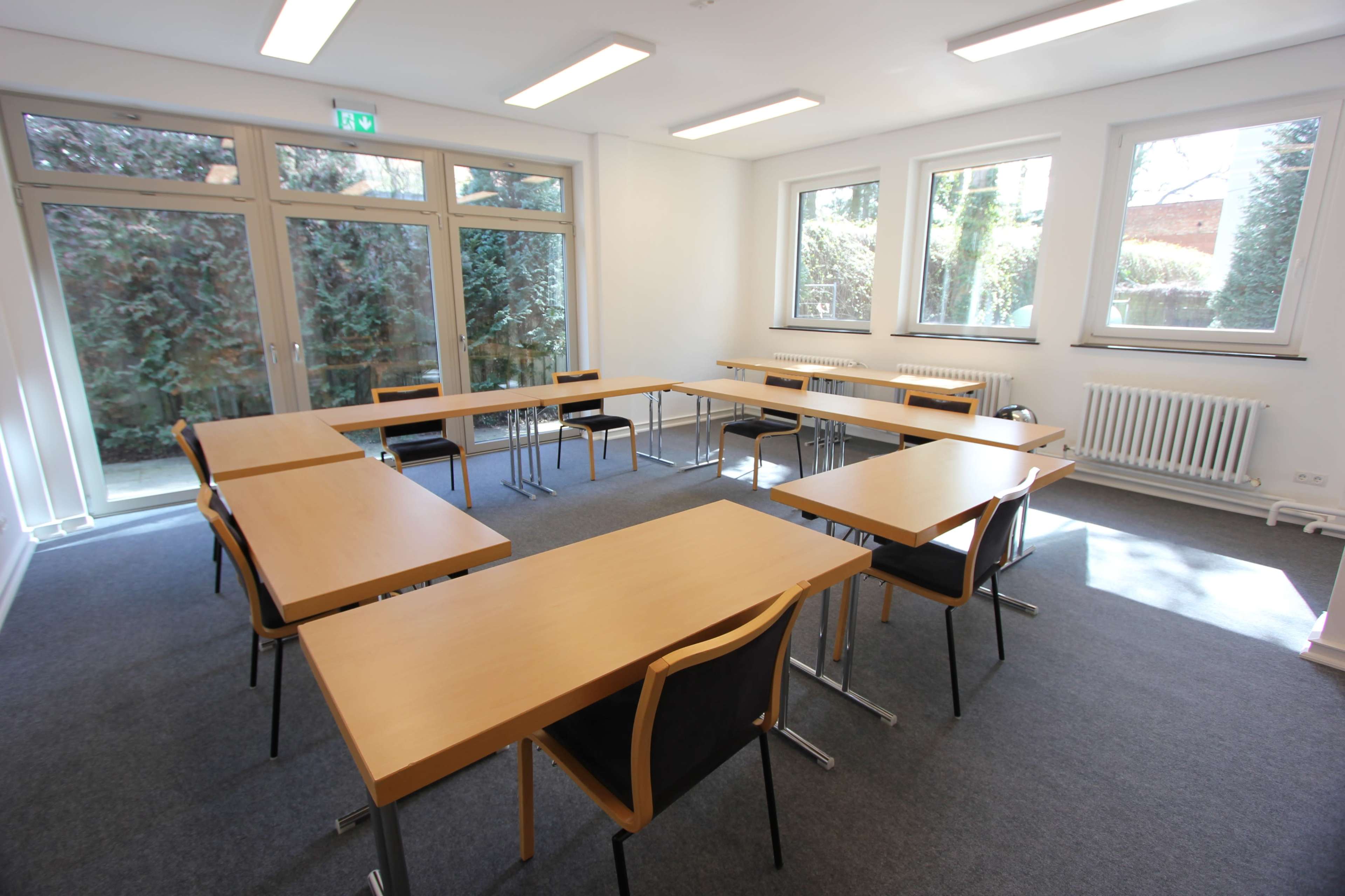 The image shows a classroom setup with several rectangular tables arranged in a U-shape, surrounded by chairs, and large windows allowing natural light.