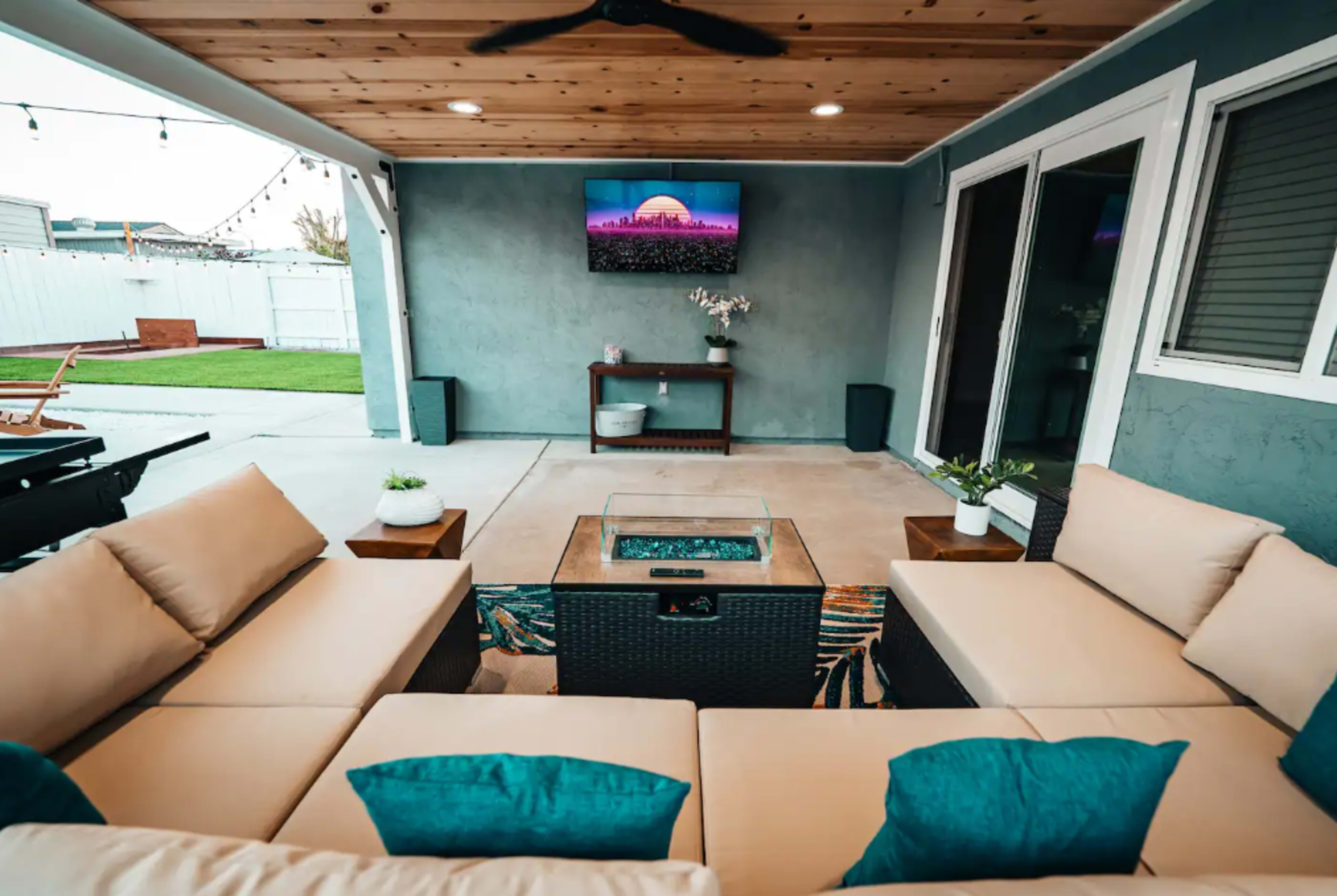 A covered outdoor living space features a sectional sofa with blue cushions, a fire pit table, and a wall-mounted television.