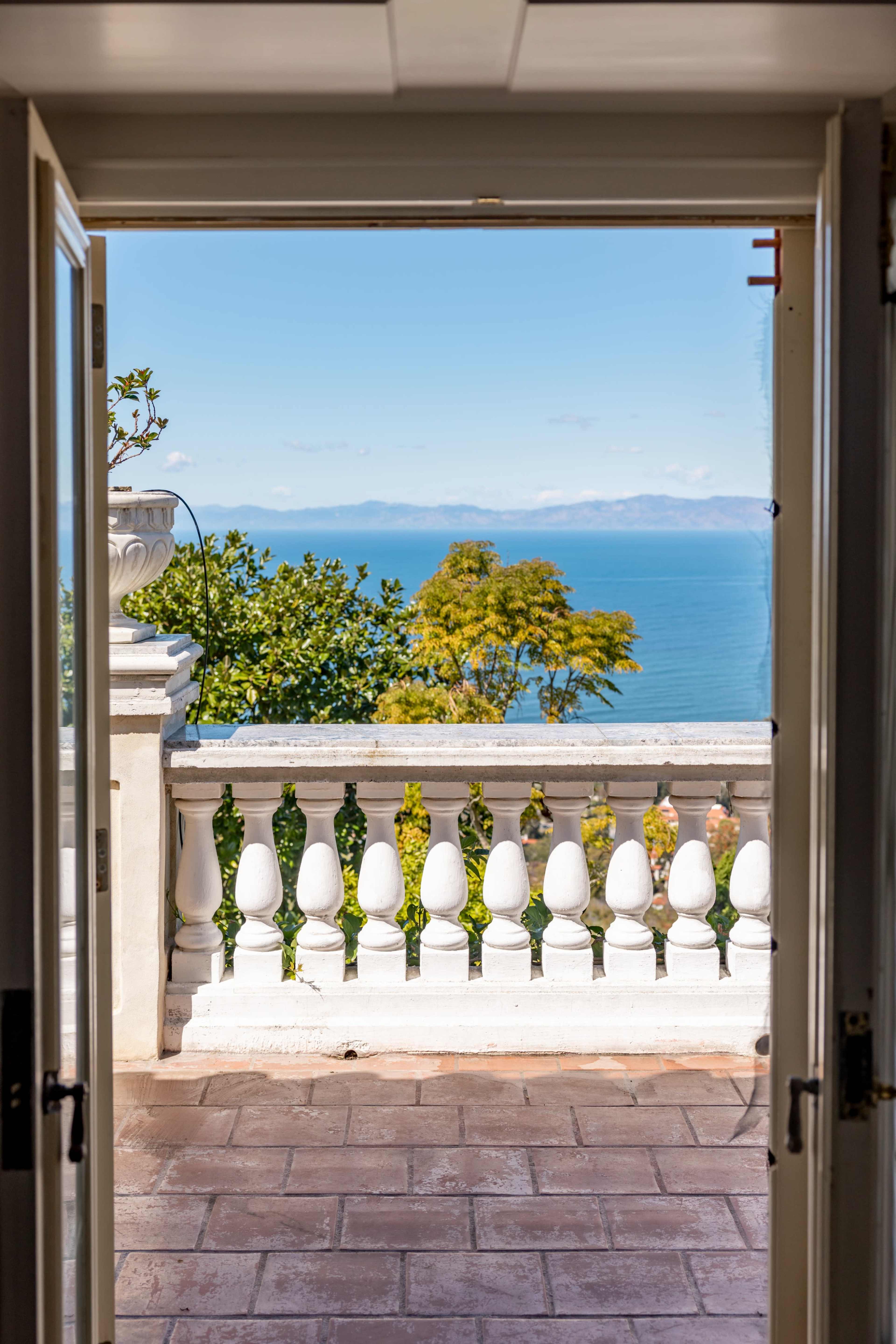 A doorway opens to reveal a view of a calm sea and distant mountains, framed by a balcony with ornate railings.