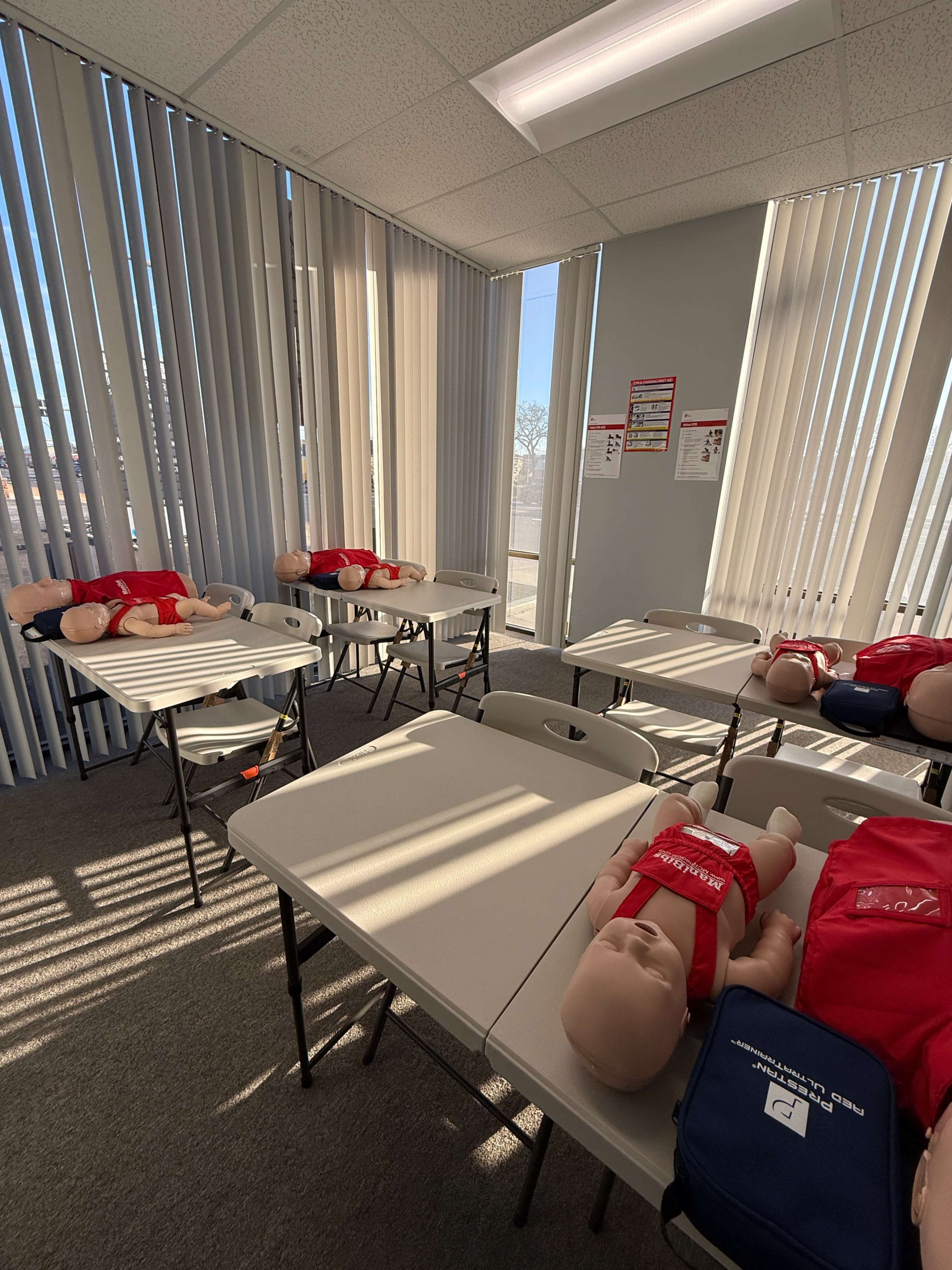 The image shows a training room with several tables, each featuring a CPR training dummy and a first aid kit in the corner.