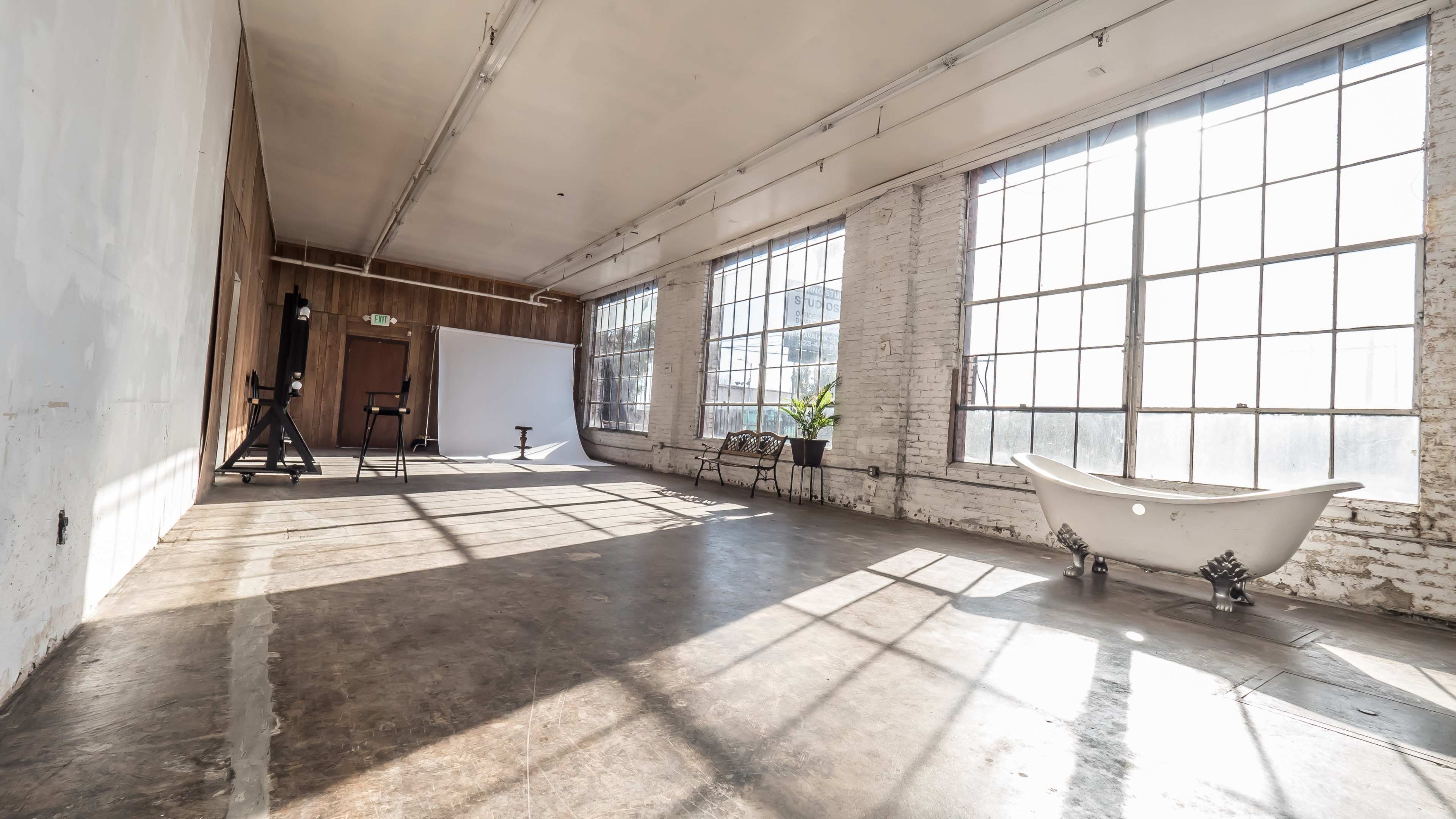 The image shows a spacious, bright studio with large windows, a clawfoot bathtub, and minimalist furnishings.