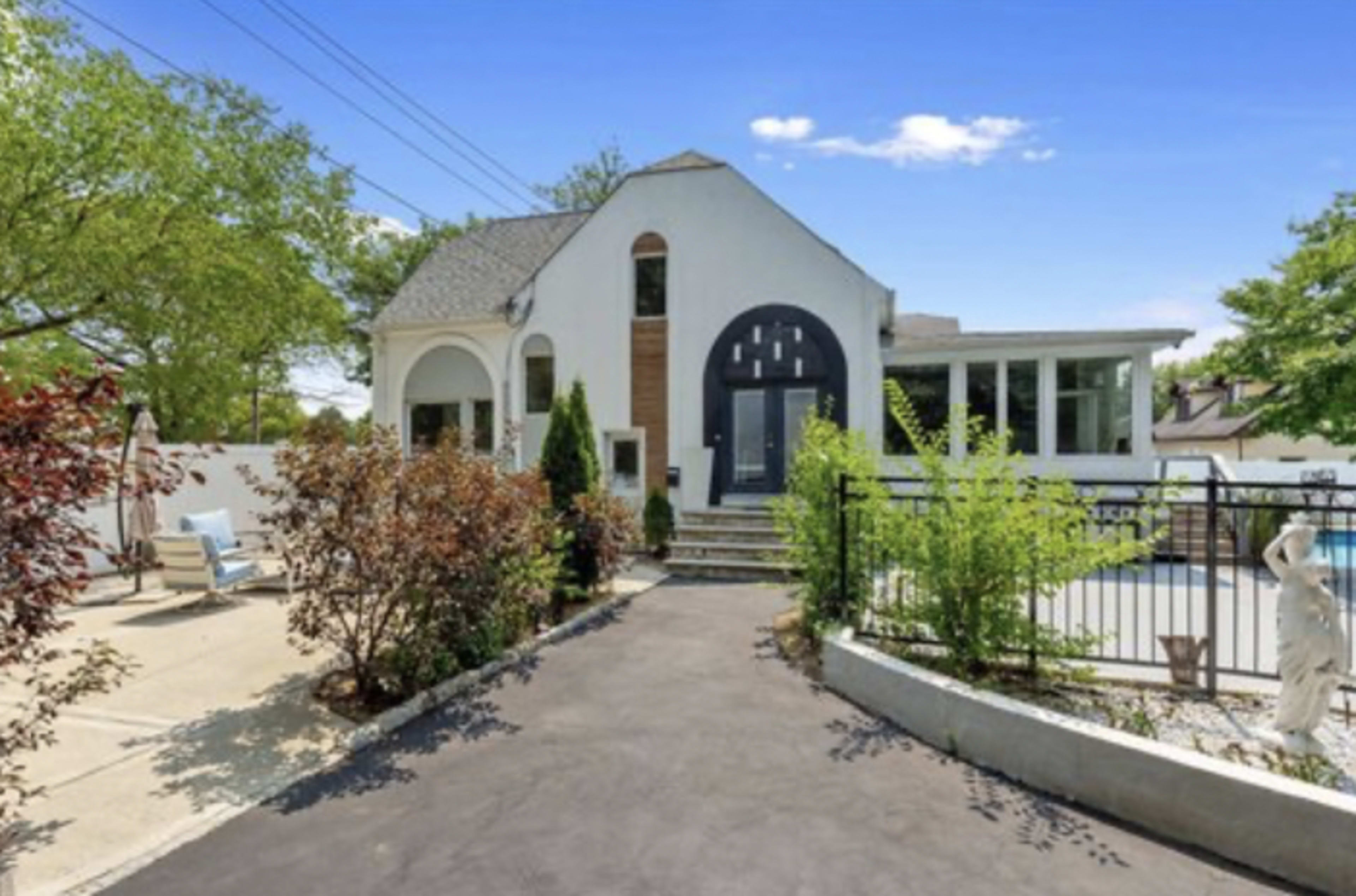 The image shows a modern house with a unique archway entrance, surrounded by greenery and a paved driveway leading to a pool area.