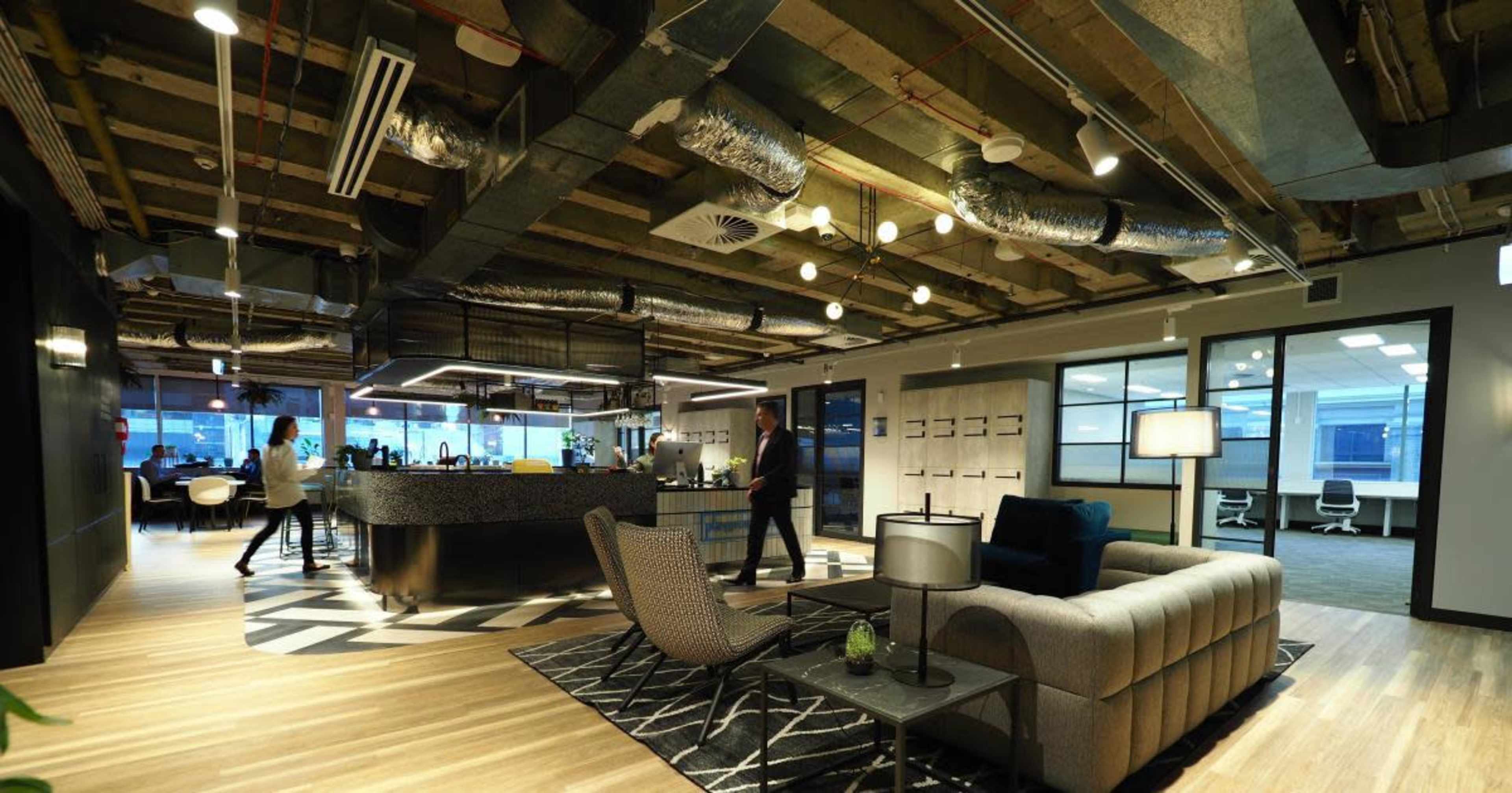 A modern office interior with an open layout, featuring a lounge area, a bar-like counter, and large windows bringing in natural light.