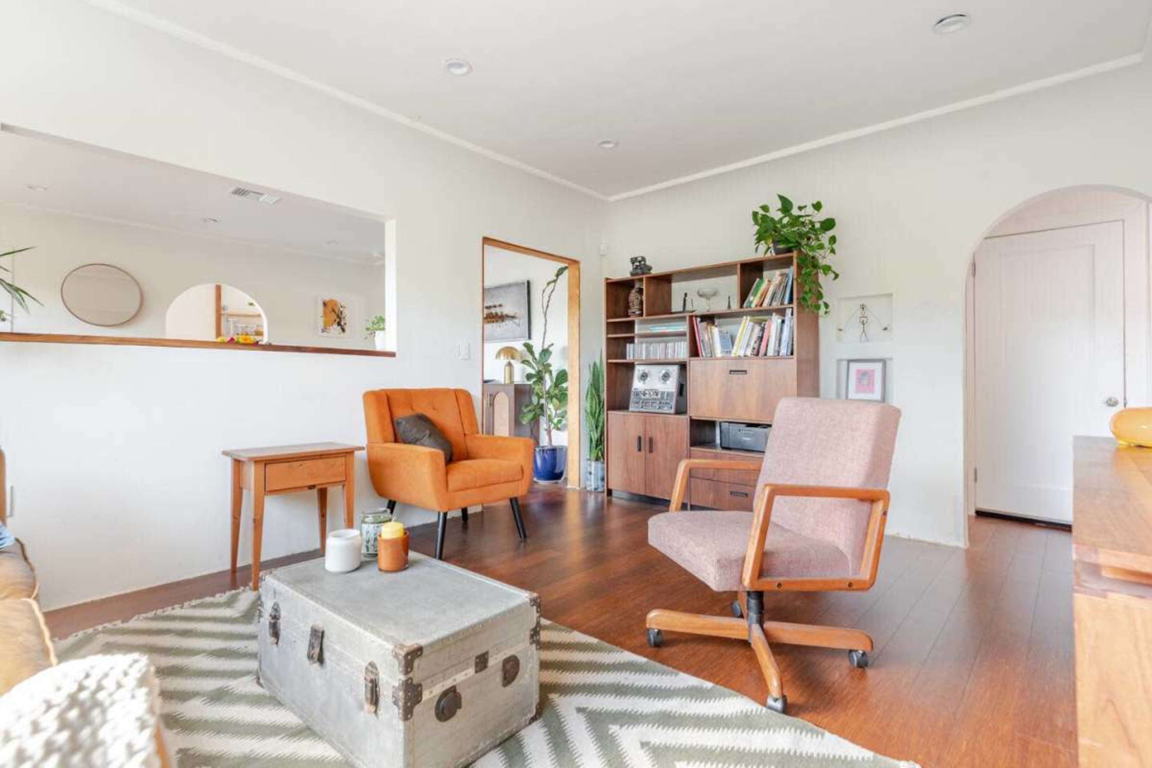 The image shows a cozy living room with a wooden coffee table, two armchairs, a bookshelf filled with books, and plants, all set on a patterned rug.