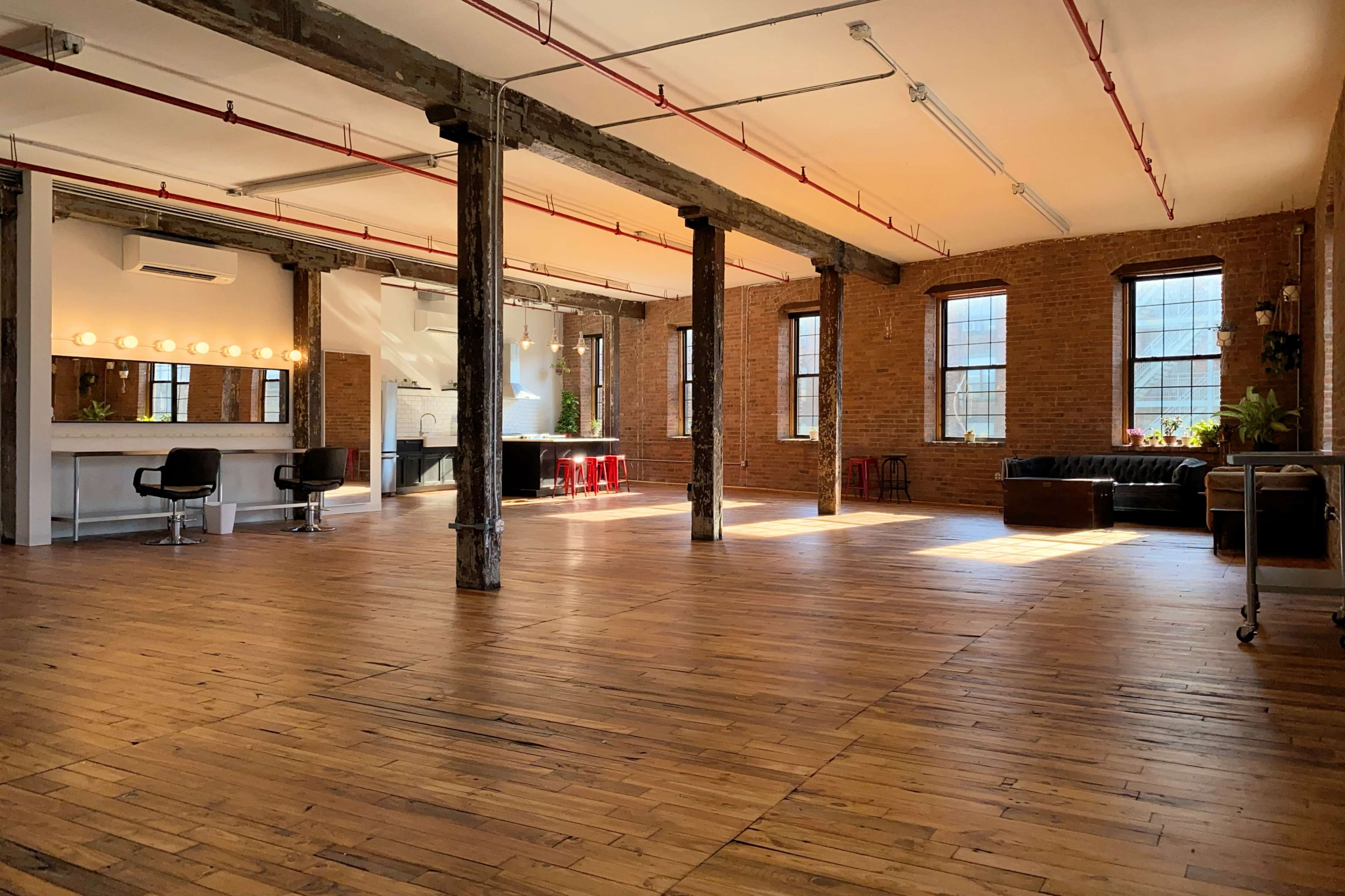 The image shows an open industrial space with exposed brick walls, wooden floors, large windows, and various seating arrangements.