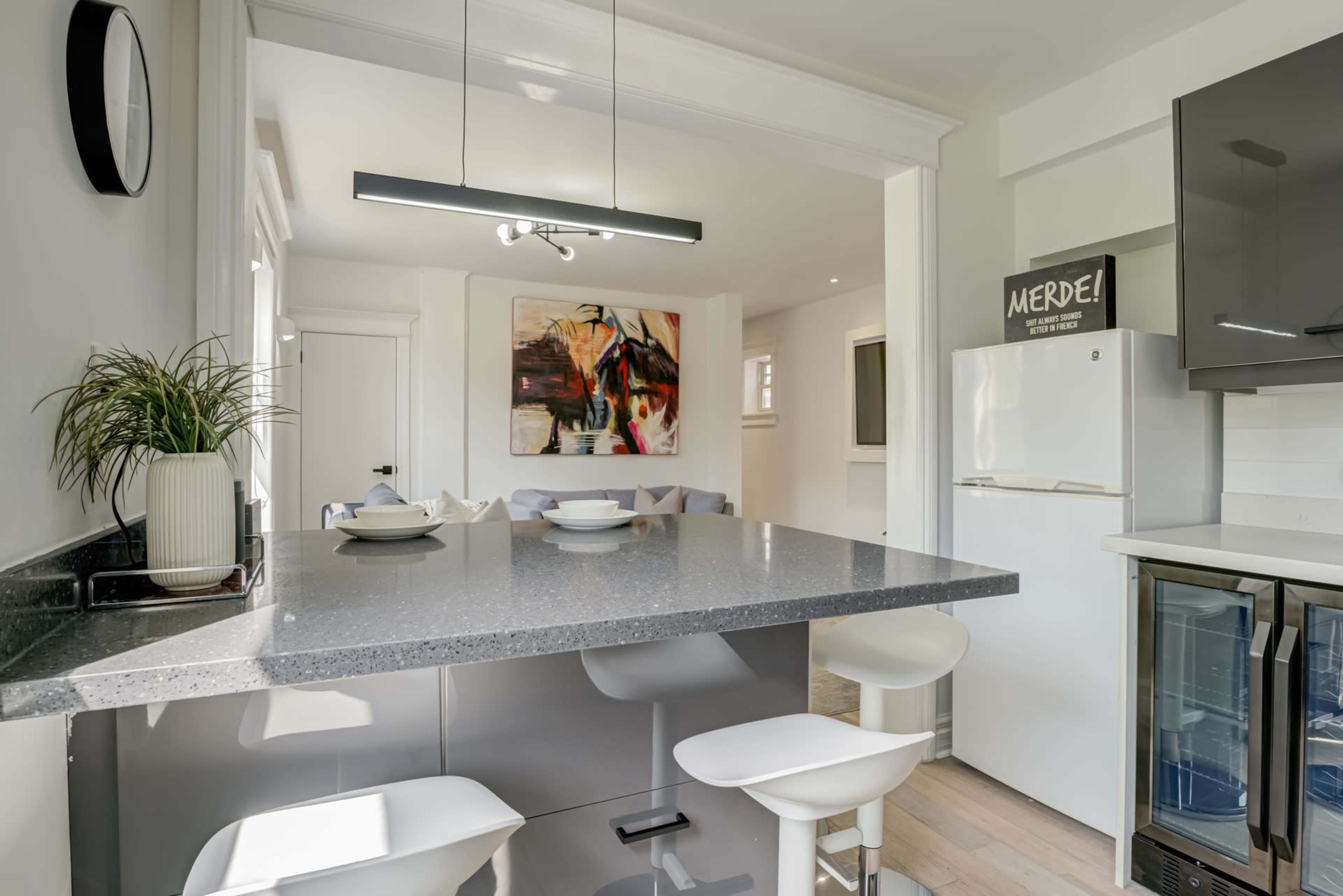 A modern kitchen features a gray island with bar stools, a white refrigerator, and a large artwork on the wall.