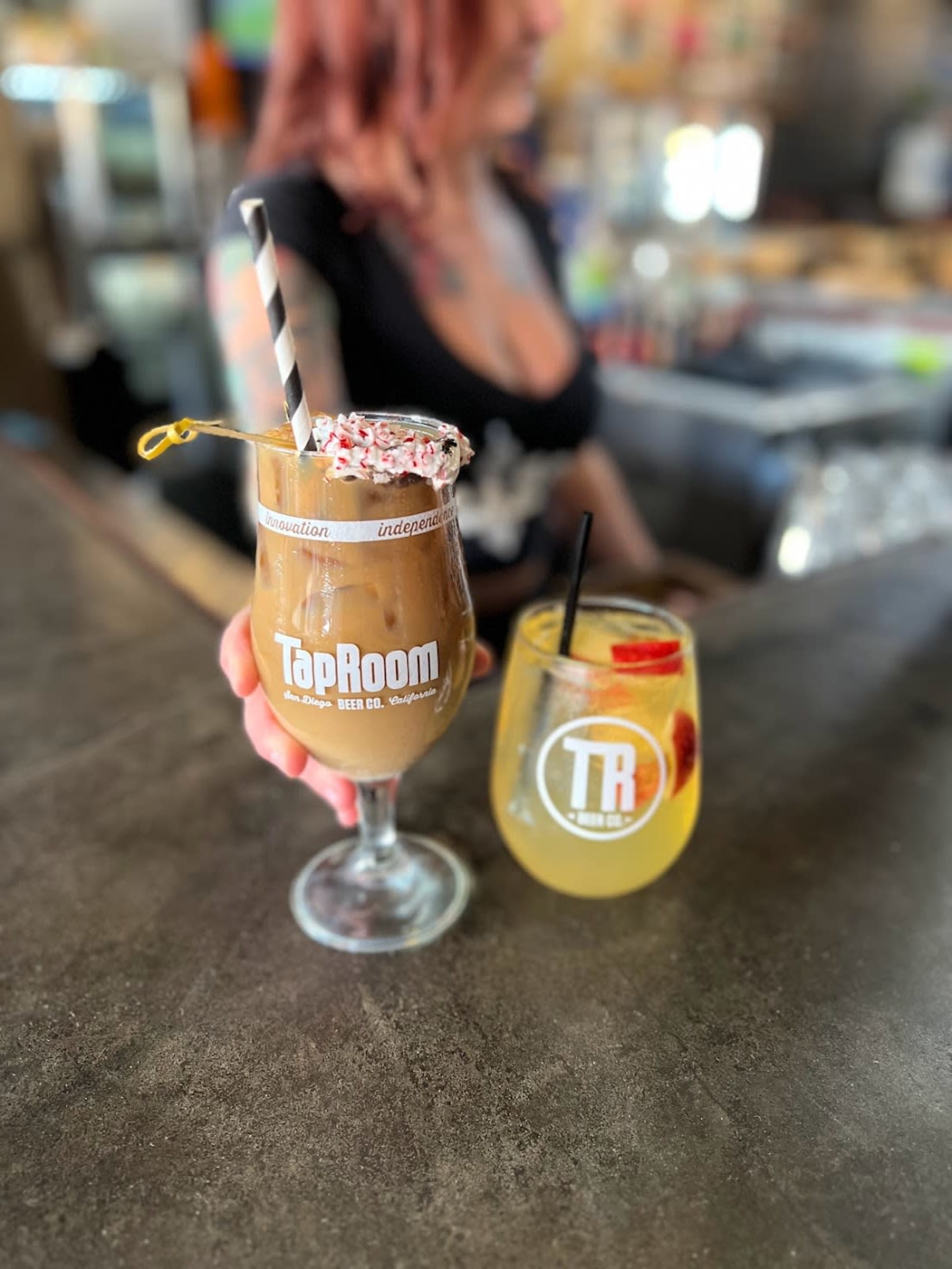 A bartender holds two colorful beverages, one in a clear glass with a straw and the other in a larger glass branded 'TapRoom.'