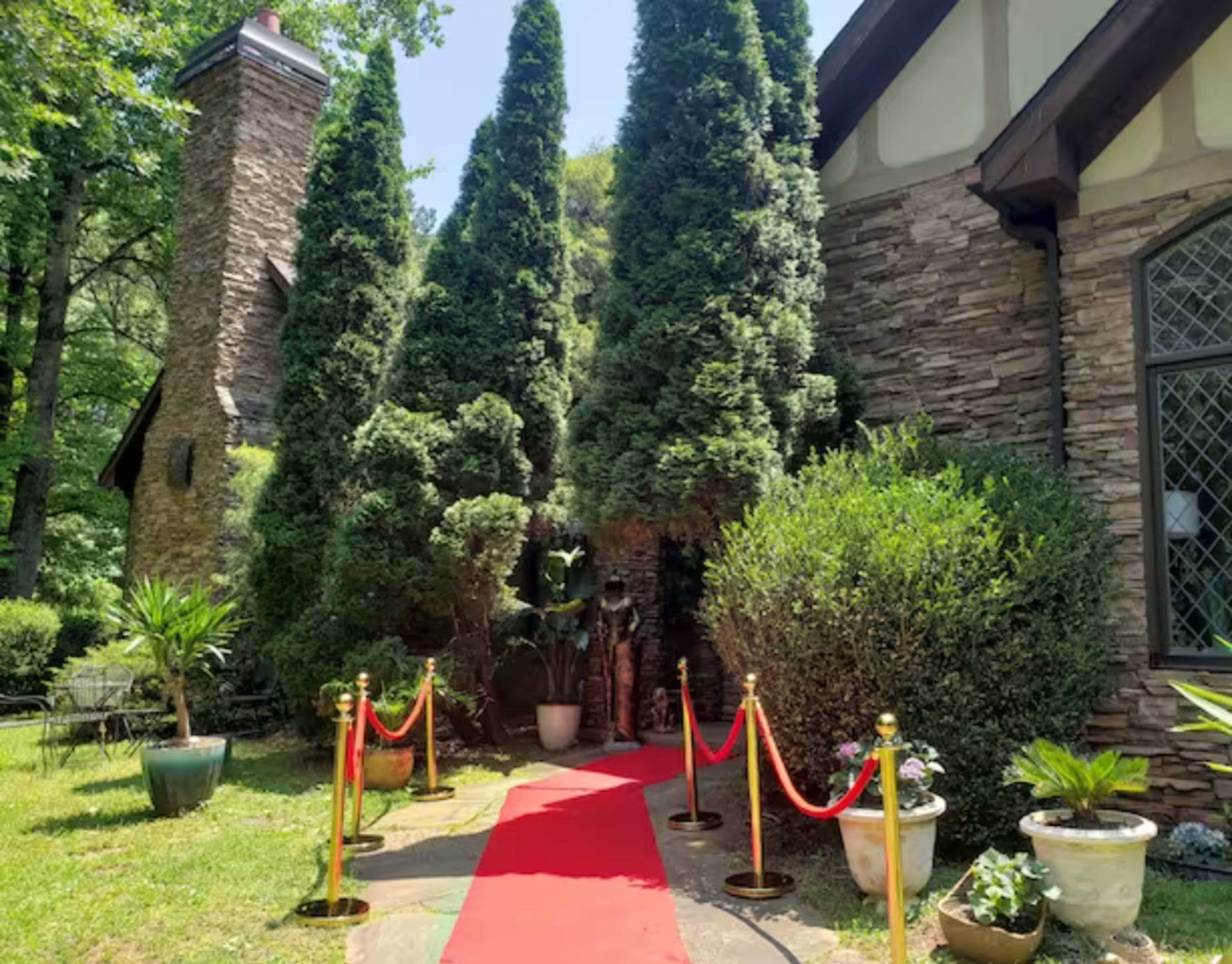 A red carpet leads to a stone building surrounded by tall conifer trees and landscaped greenery.
