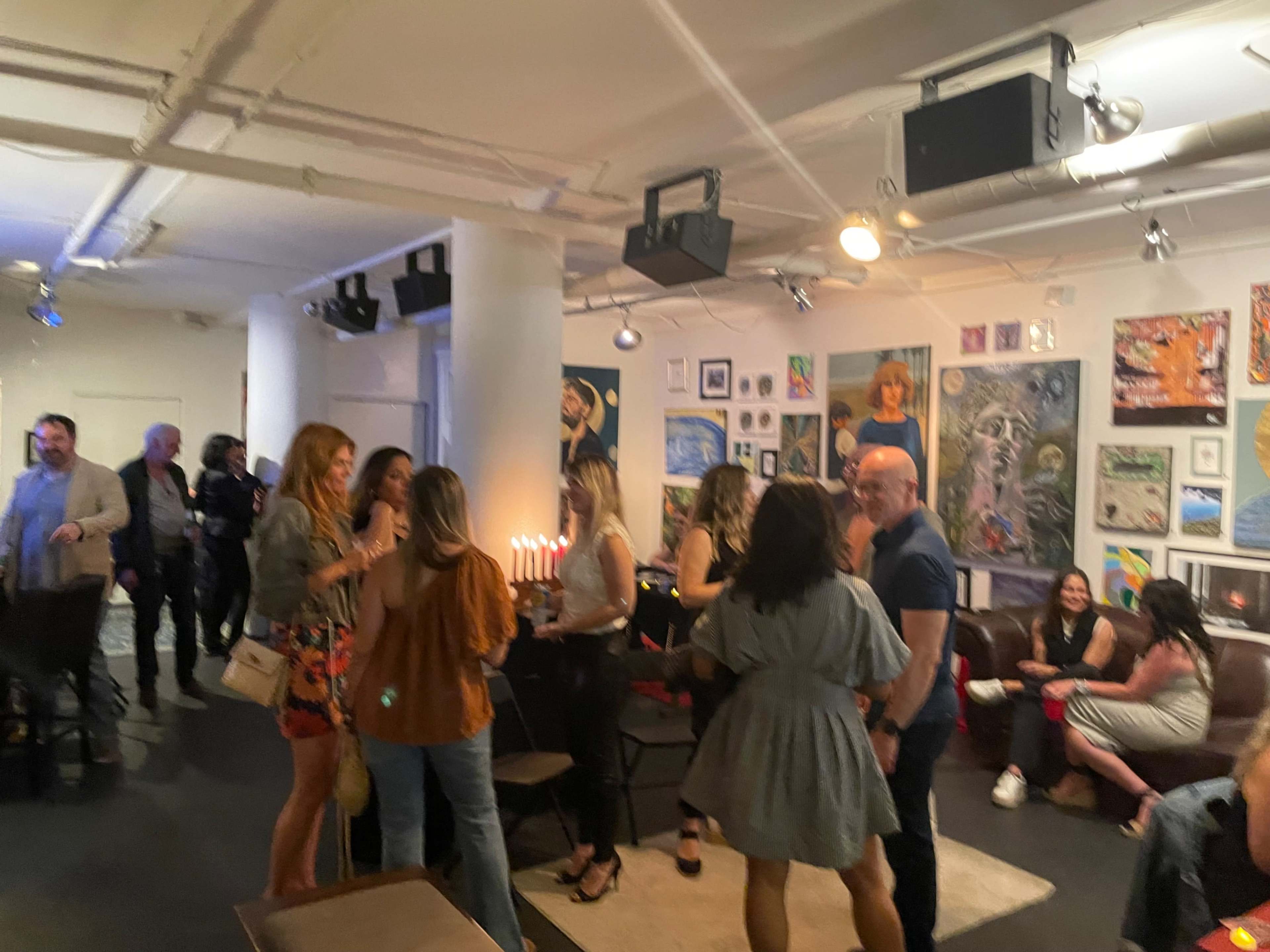 A group of people socialize in an art-filled venue with various paintings on the walls and soft lighting.