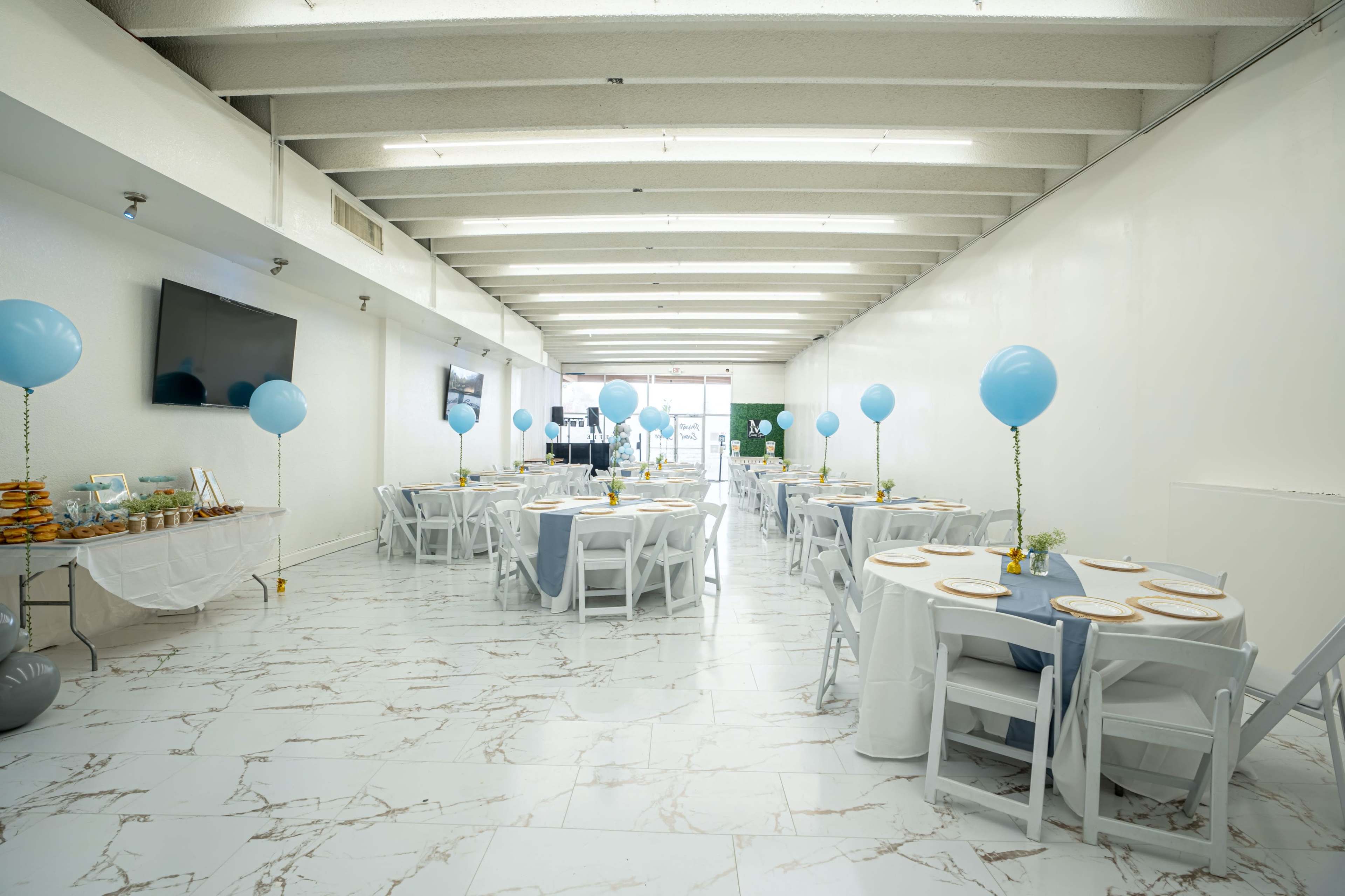 A spacious event hall features neatly arranged tables with blue balloons and decorative centerpieces.