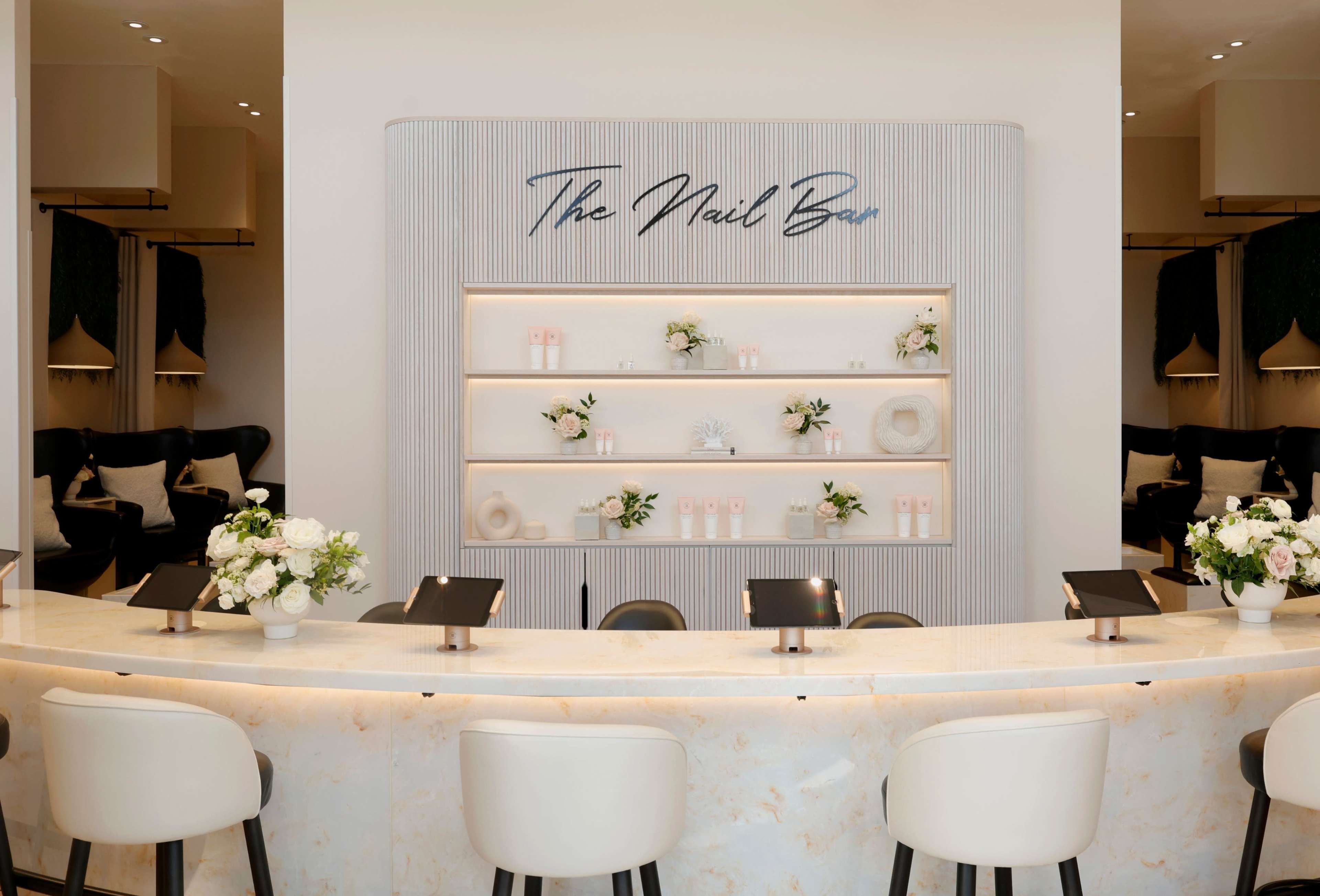 The image shows a neatly arranged nail bar reception area with a marble countertop, backlit shelves displaying products, and elegant floral arrangements.