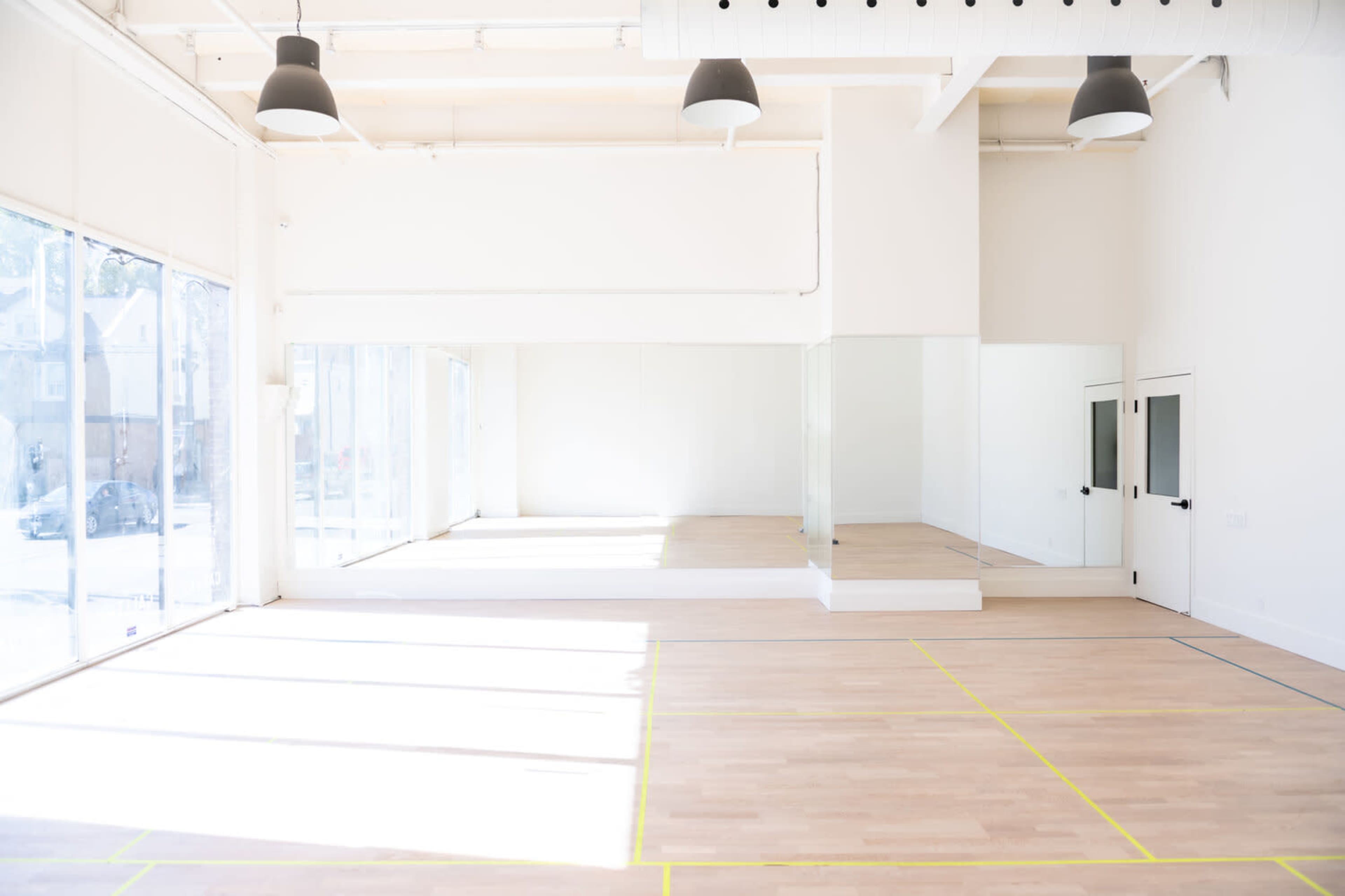 A bright, empty studio space with wooden floors, large windows, and a mirrored wall.