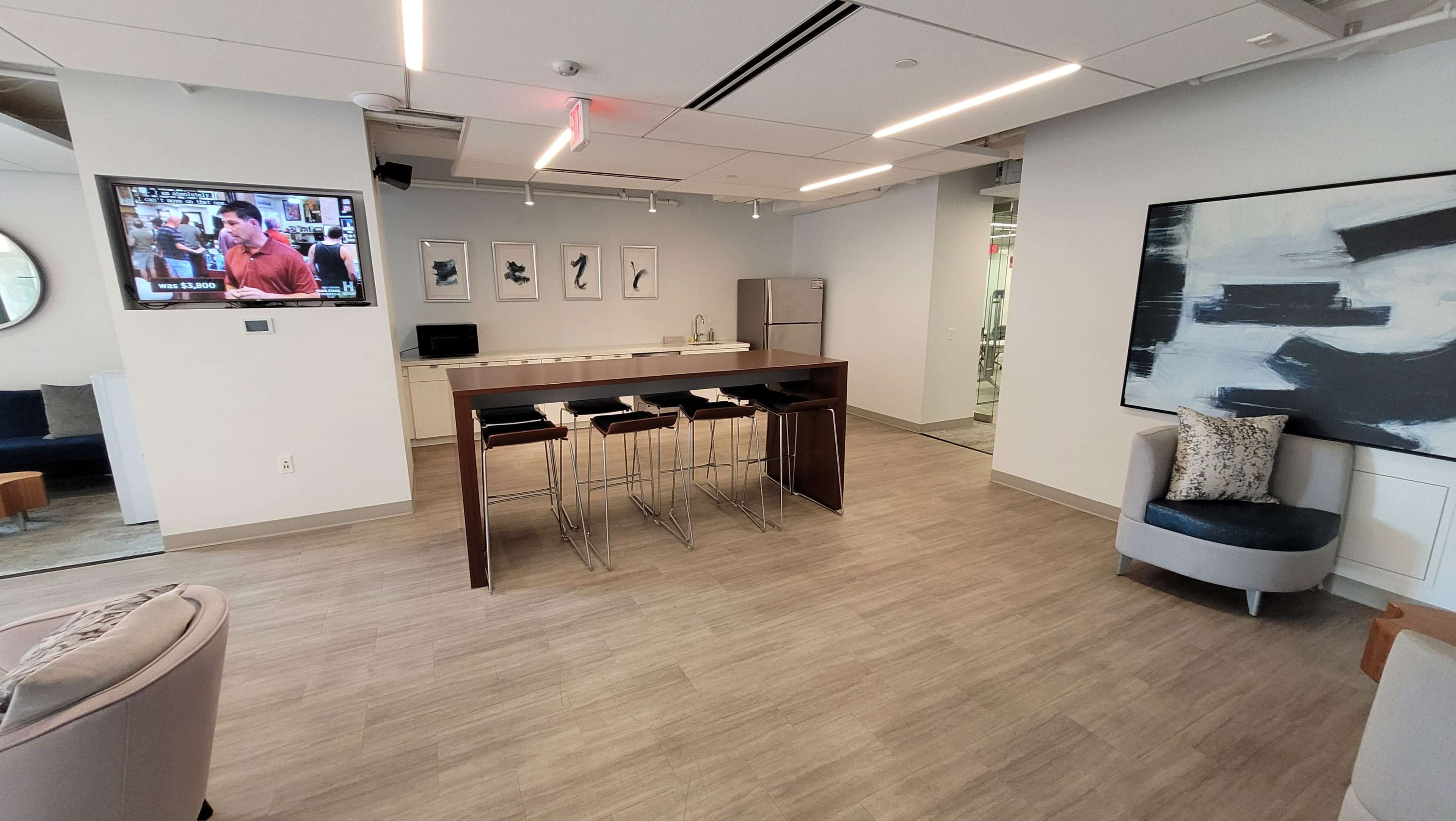 The image shows a modern lounge area with a kitchen counter, bar stools, and a television mounted on the wall.