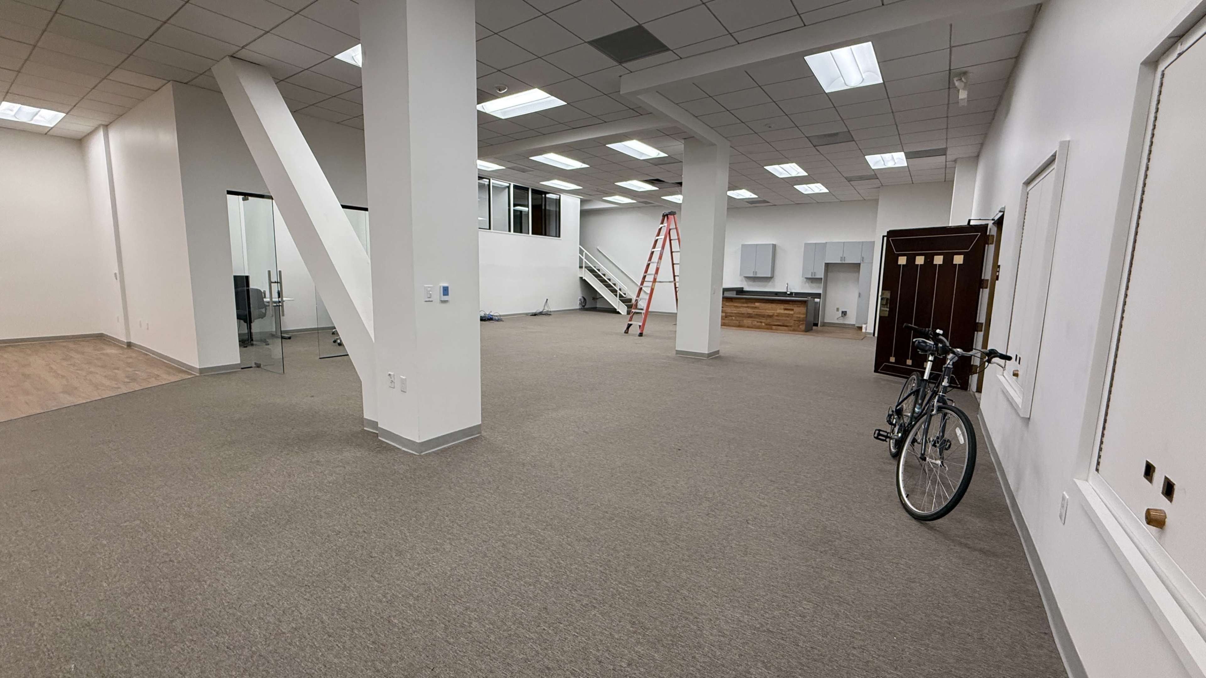 The image shows a spacious, empty office interior with beige carpeting, large windows, a kitchen area, a ladder, and a bicycle parked against the wall.