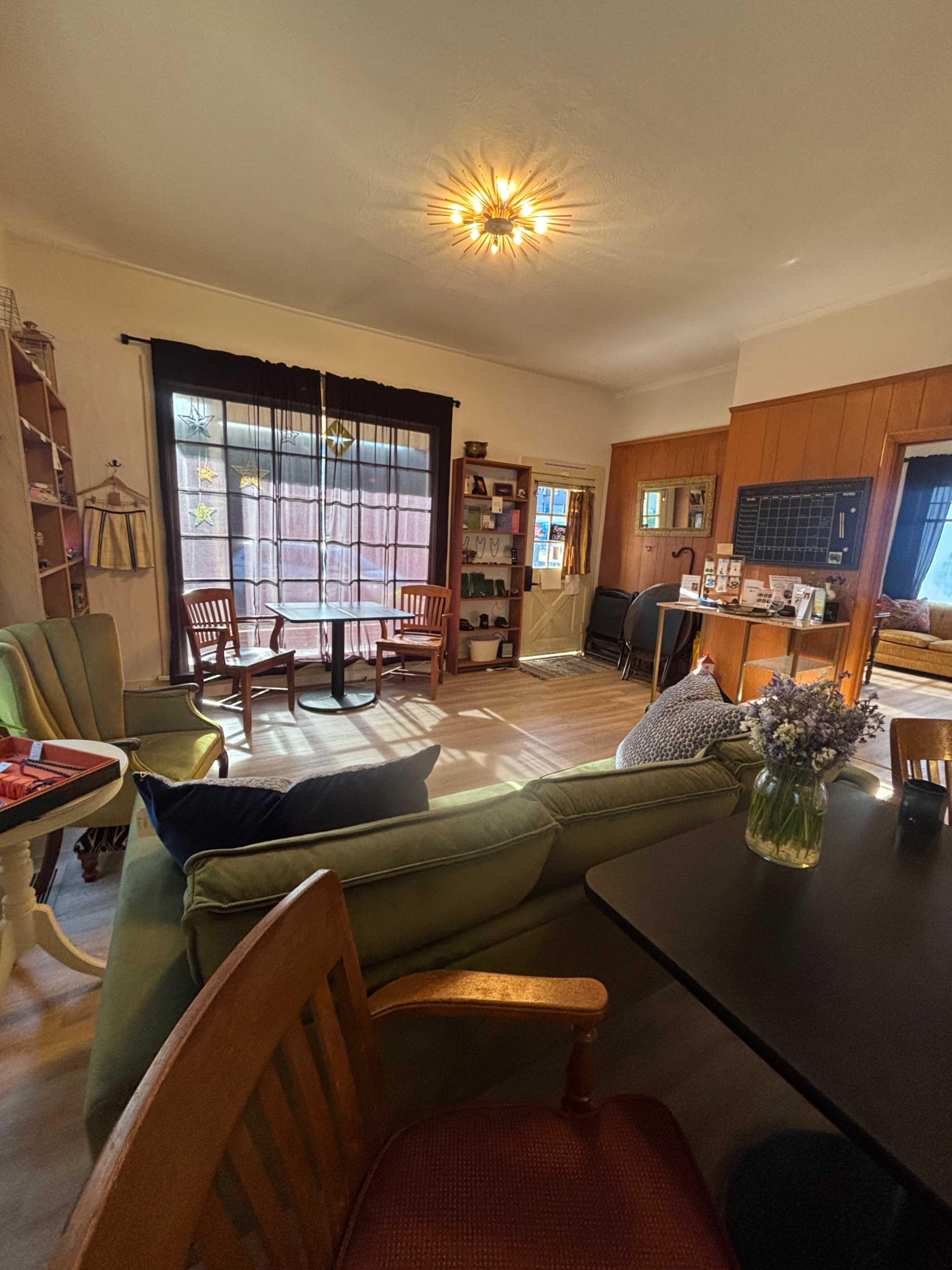 The image shows a cozy living space with green and brown furniture, a bright window with sheer curtains, and various decorative elements on the walls and shelves.