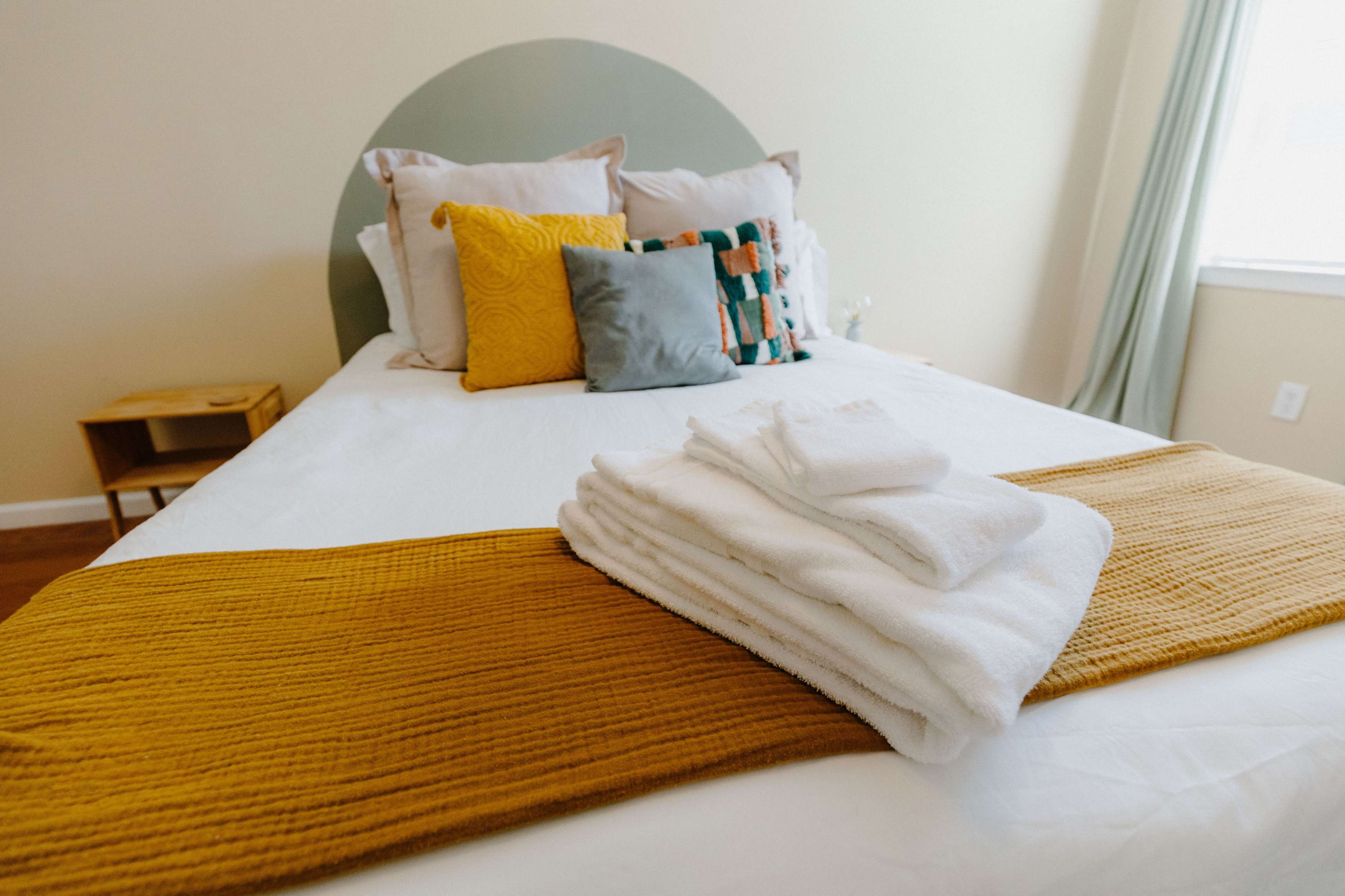 A neatly made bed features folded white towels and a mustard-colored blanket, with decorative pillows arranged on the headboard.