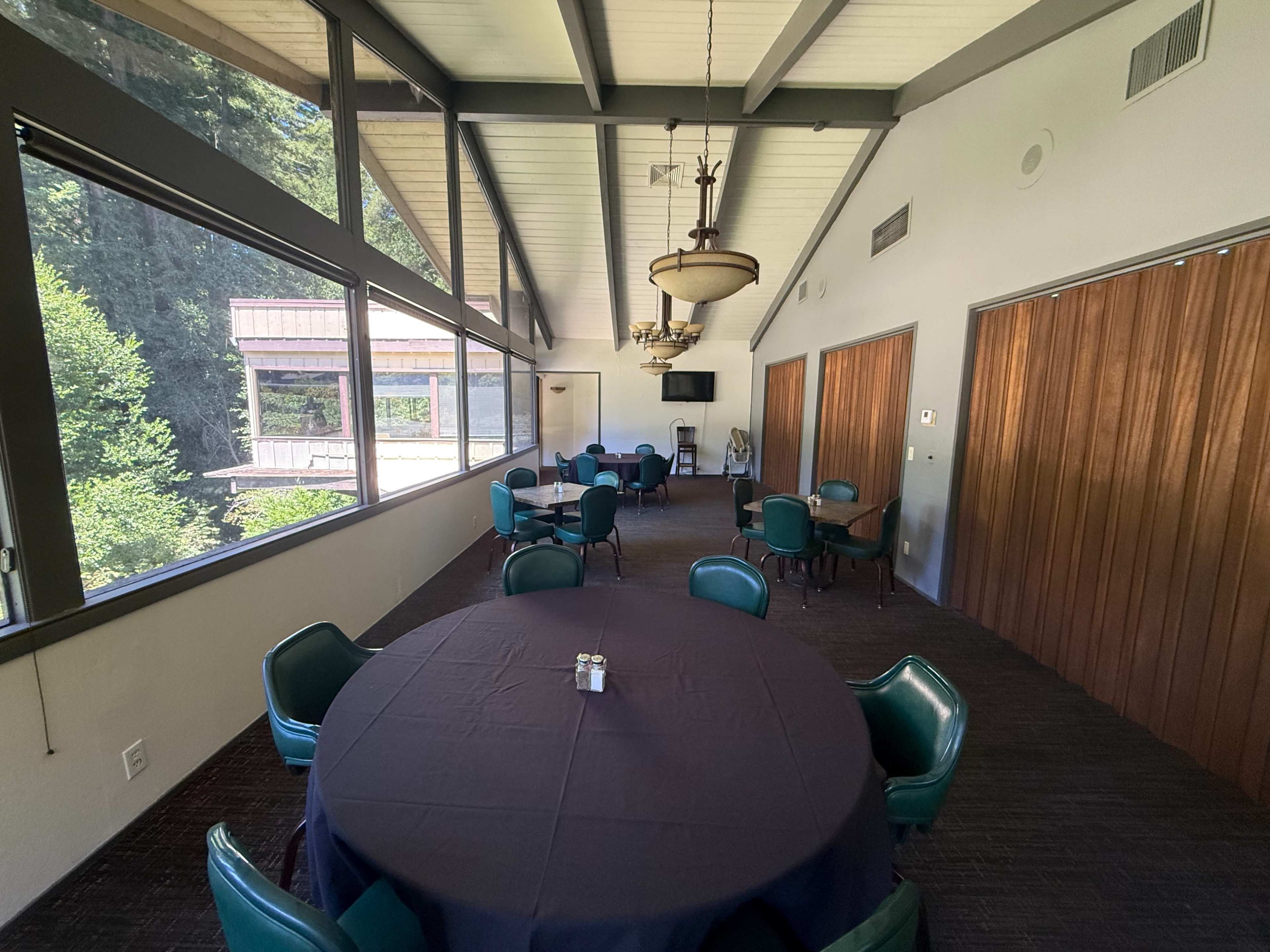 The image shows a spacious indoor room with large windows, featuring several round tables covered with dark cloth and chairs arranged around them.
