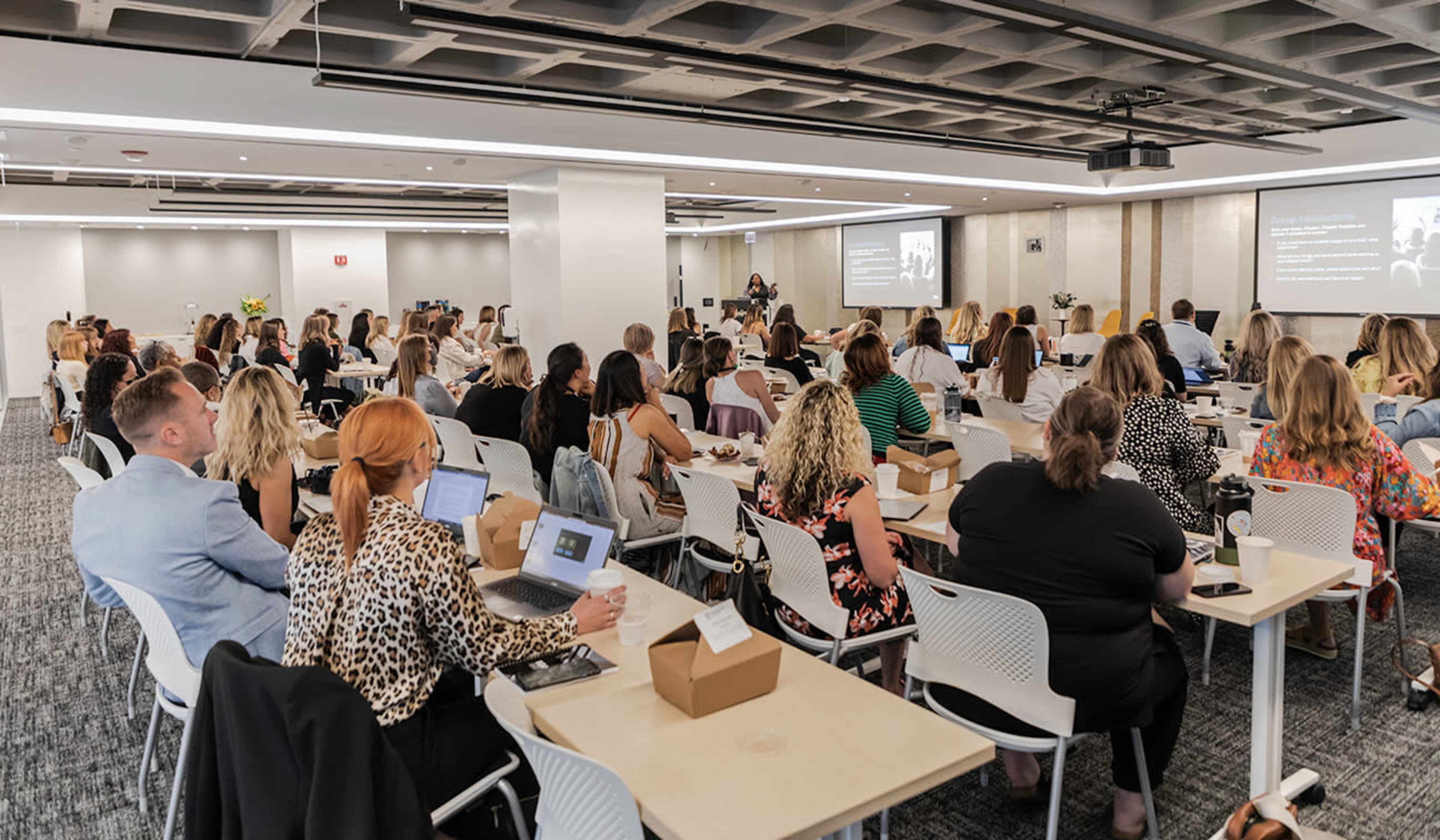 A group of attendees sits at tables in a well-lit conference room, focusing on a speaker presenting at the front.
