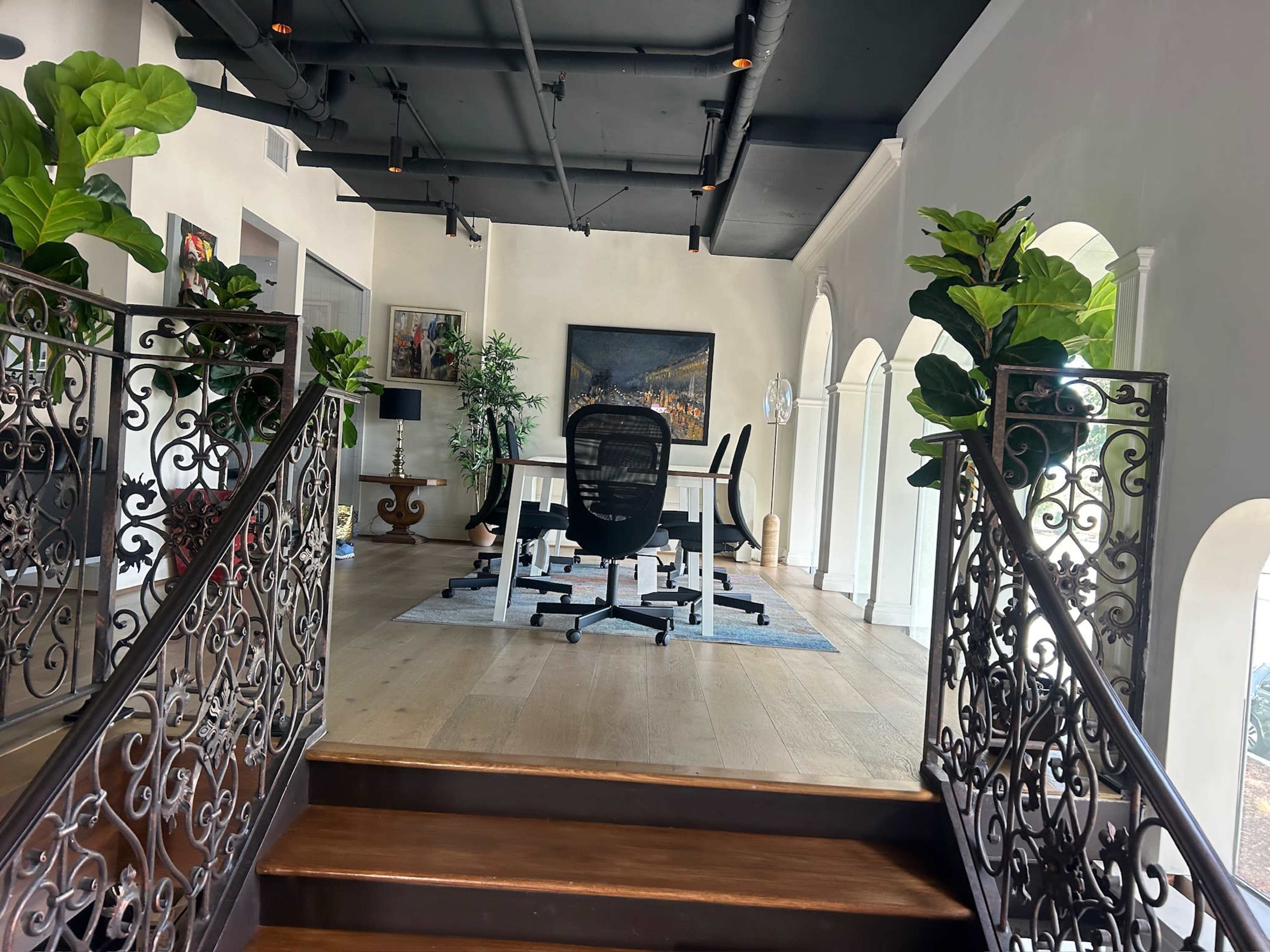 The image shows a spacious office area featuring a white desk and black office chairs, surrounded by large plants and framed artwork, with a decorative staircase leading to the entrance.