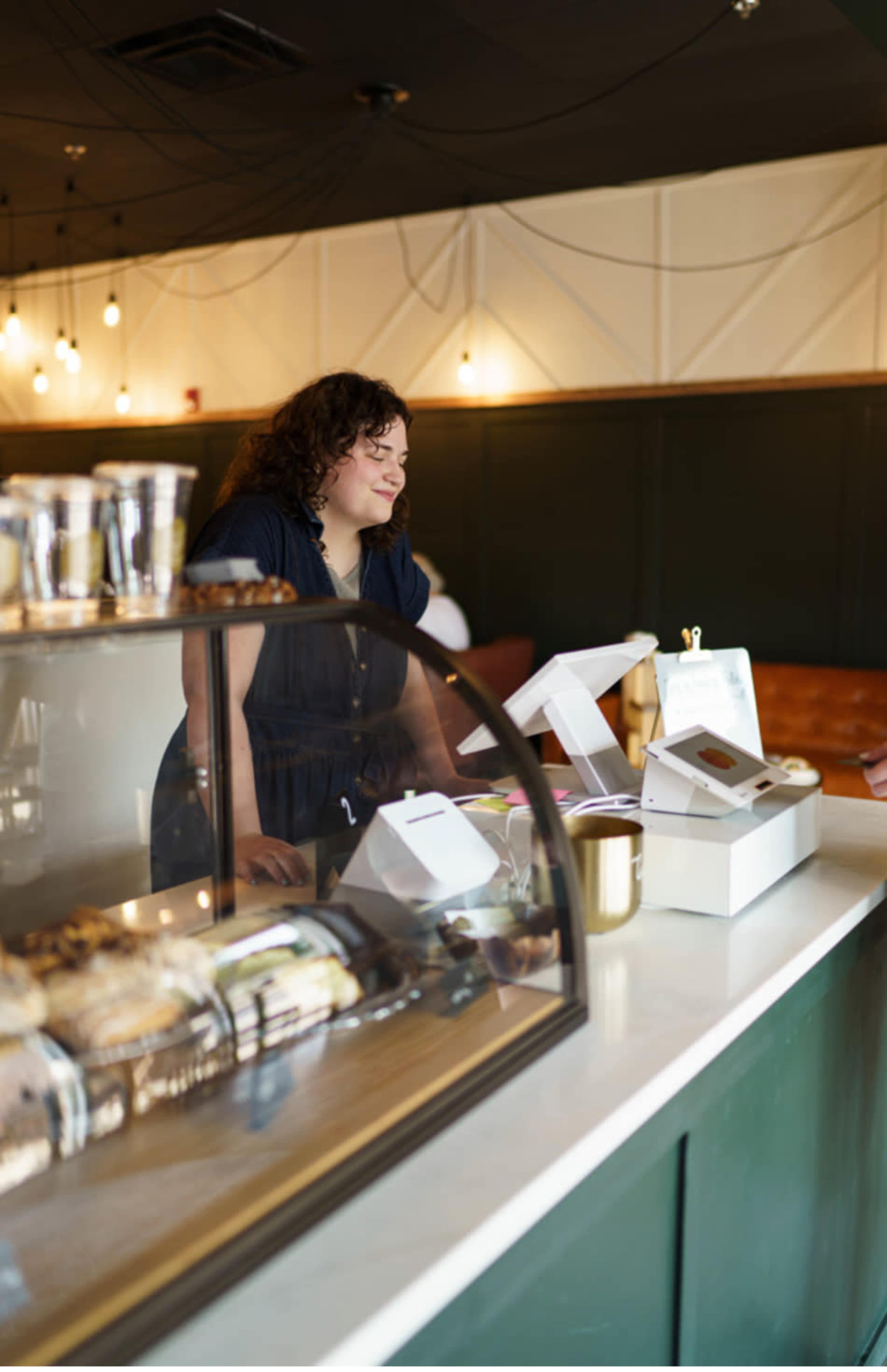 A barista stands behind a counter in a café, engaging with customers while a display case of pastries is visible in the foreground.
