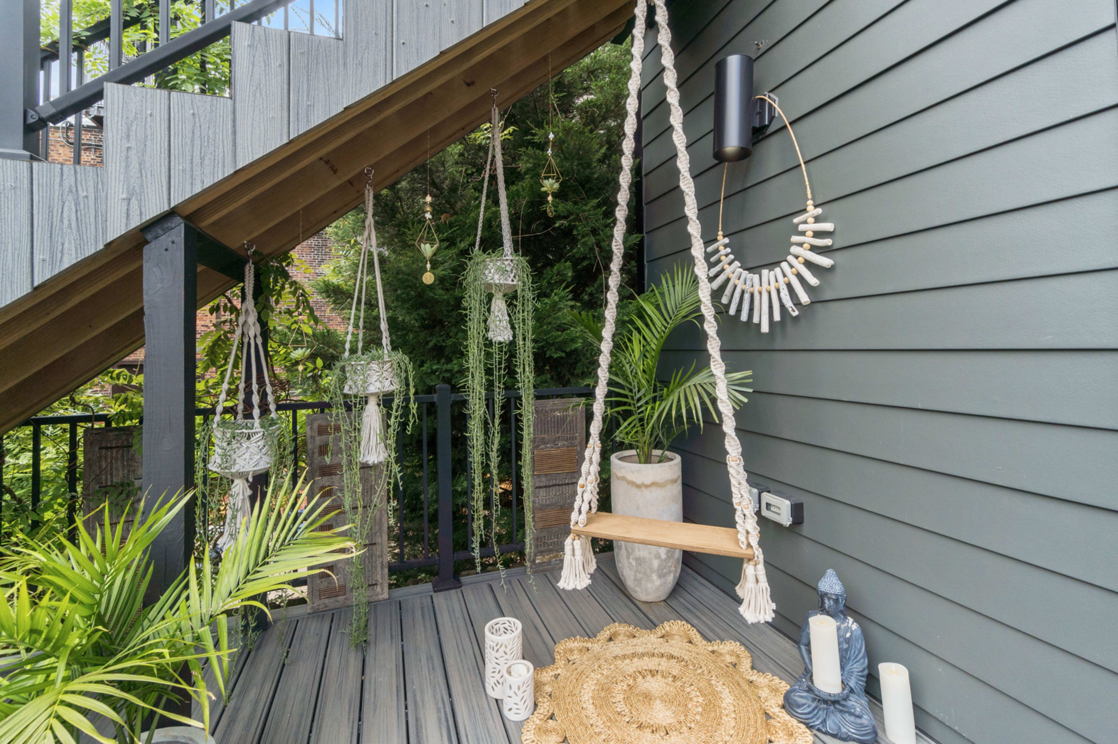 A wooden swing hangs from a staircase, surrounded by decorative hanging planters, greenery, and a stone statue.
