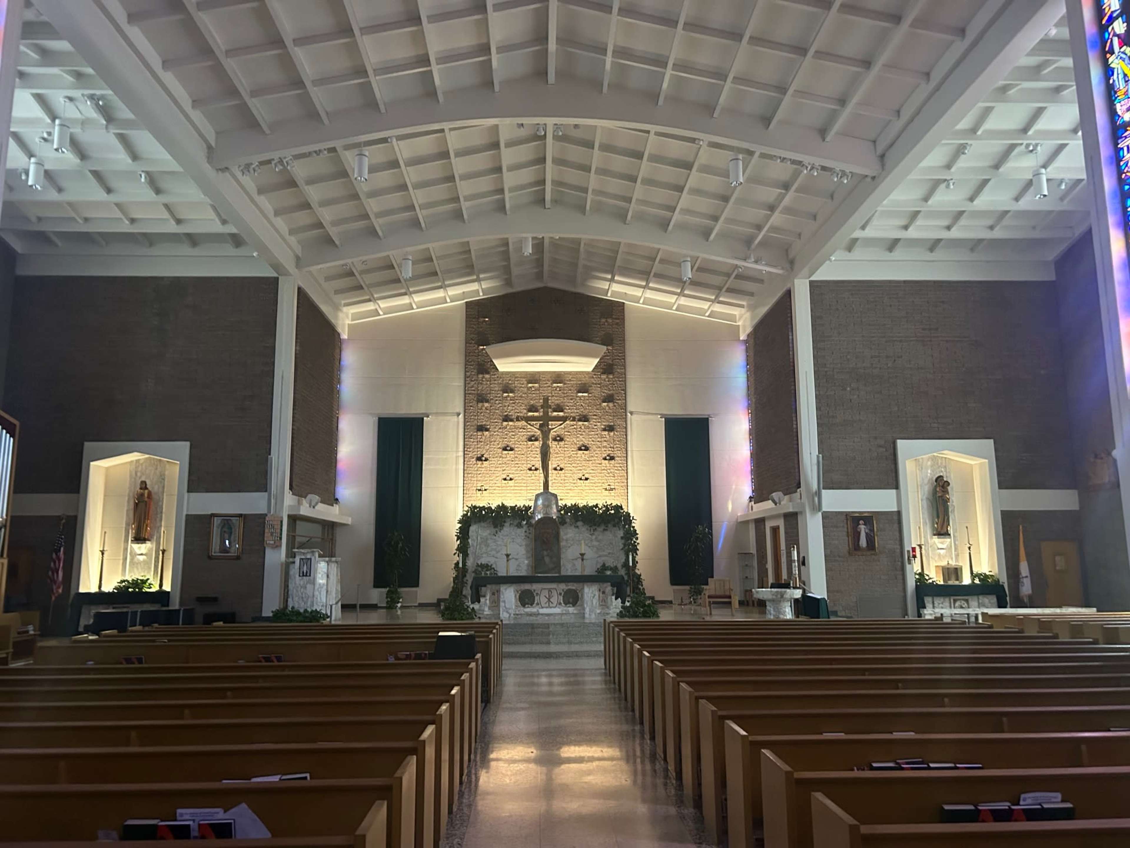 The interior of a church features a large altar with a cross, flanked by two illuminated recesses, and rows of wooden pews facing the front.