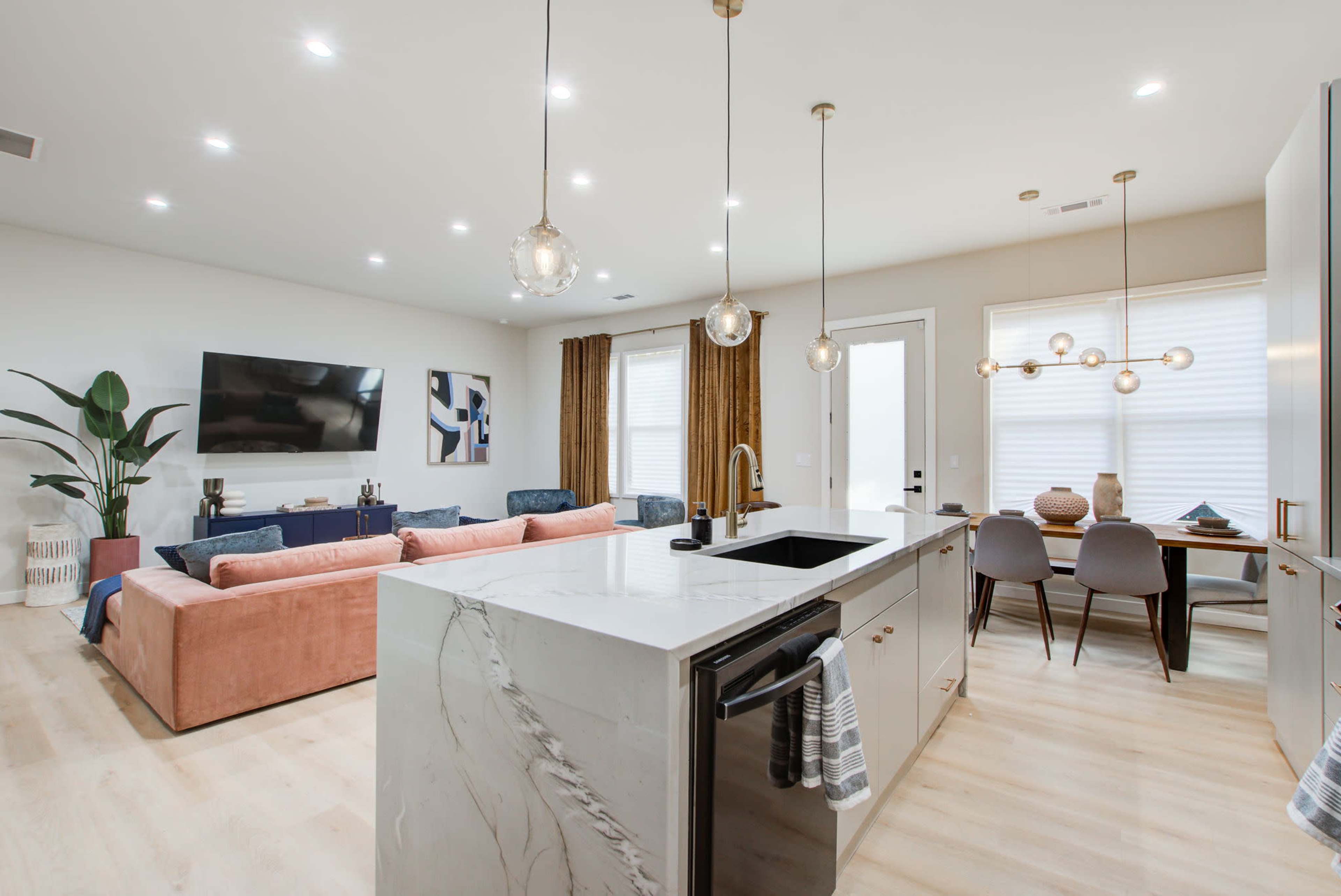 The image shows a modern living space featuring a kitchen island with pendant lights, a seating area with a sofa, and a dining table beside large windows.