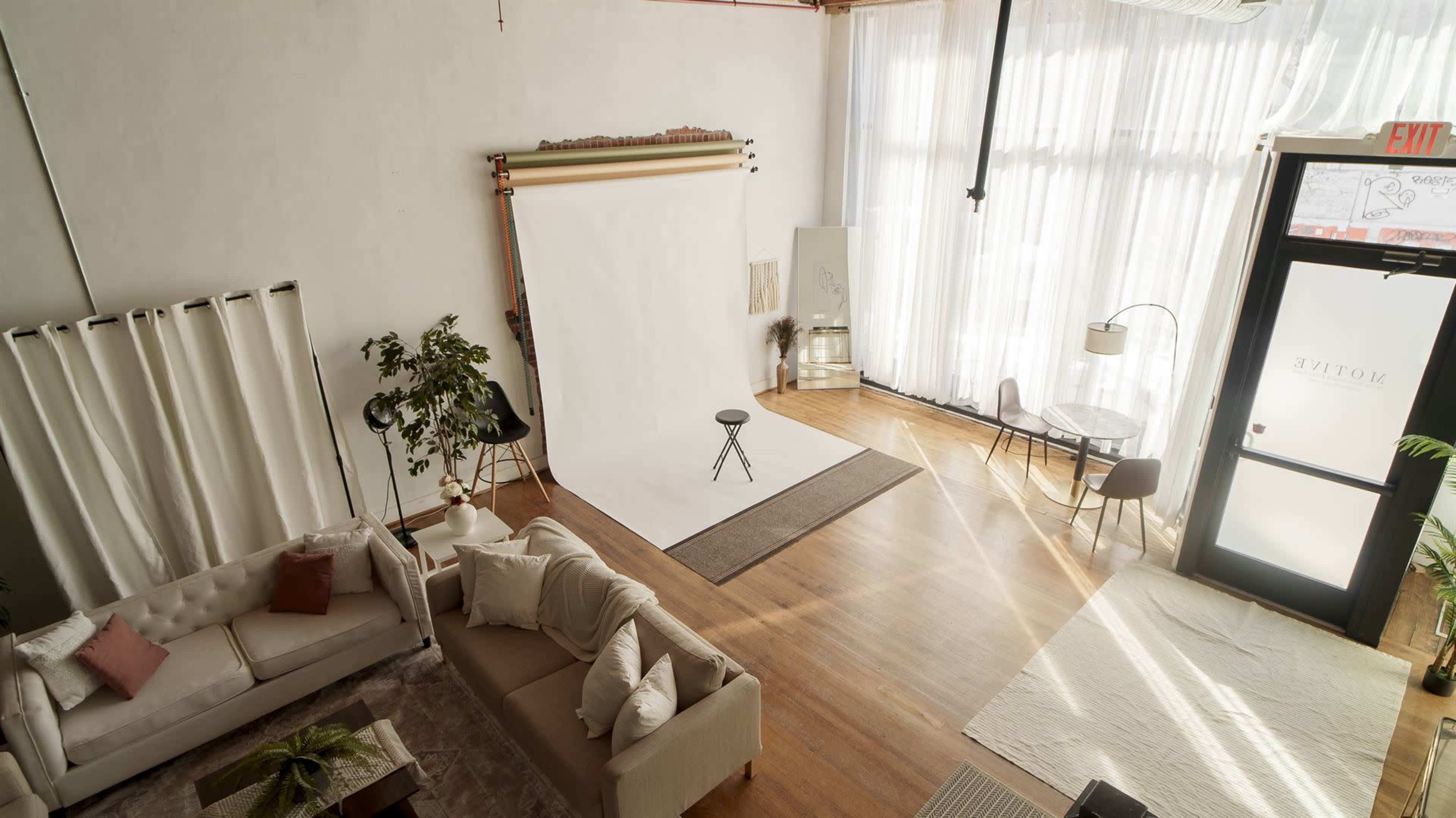 The image shows a well-lit interior photography studio featuring a white backdrop, a stool, and a comfortable seating area with a table.