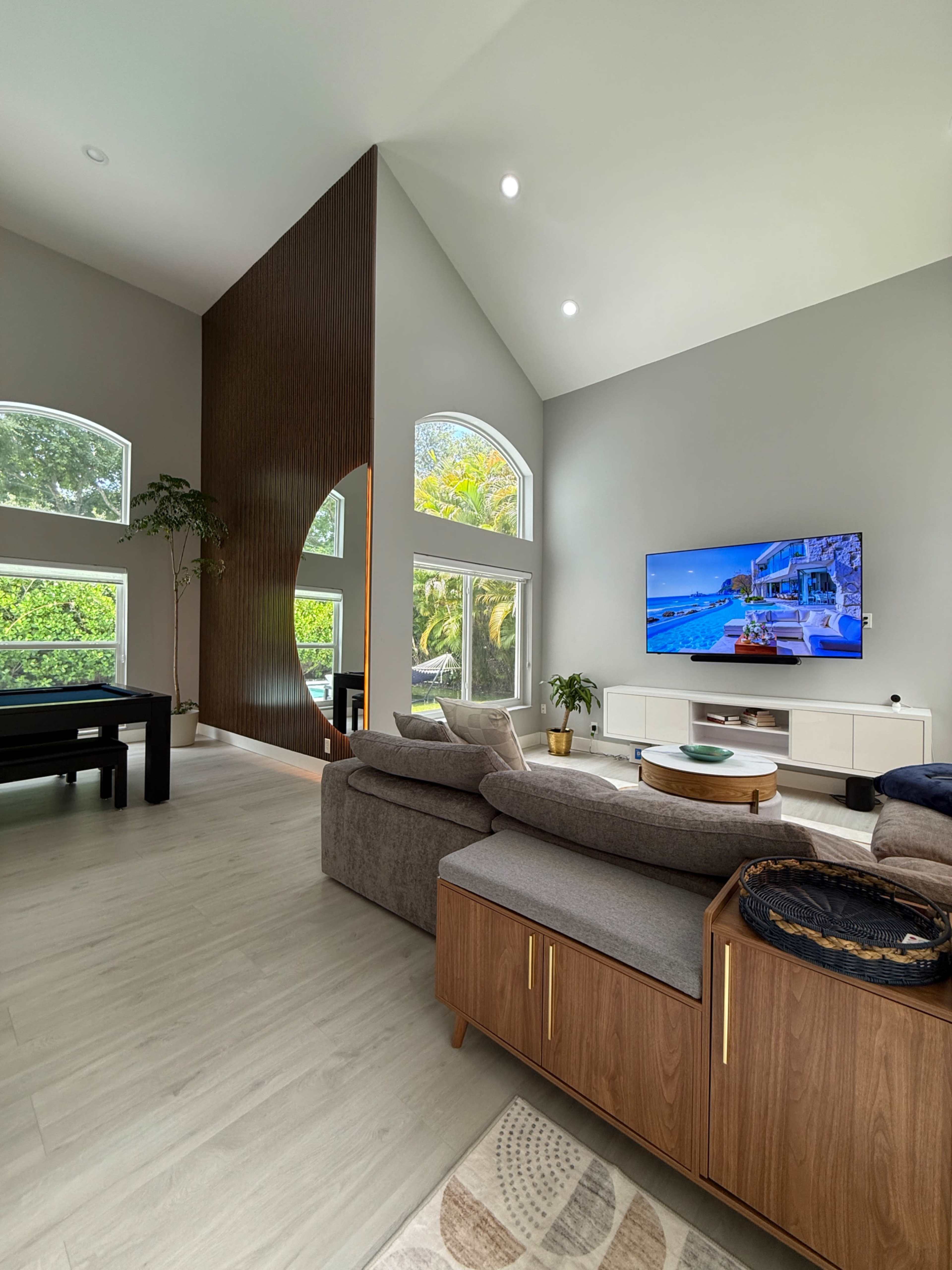 The image shows a modern living room featuring a high ceiling, large windows, a piano, a sectional sofa, and a television mounted on the wall.