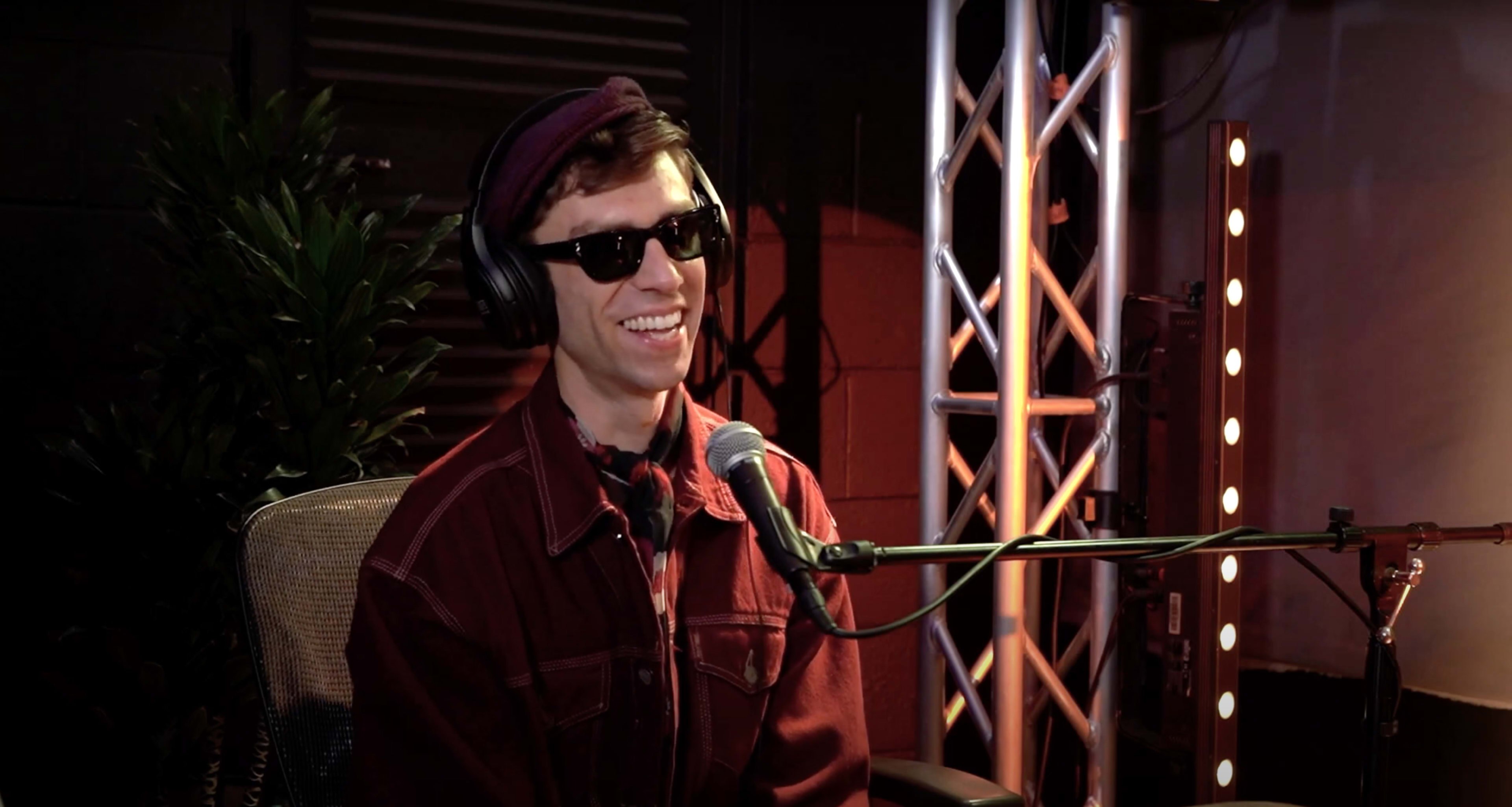 A man wearing sunglasses and headphones sits in front of a microphone, surrounded by lighting rigs and a plant.