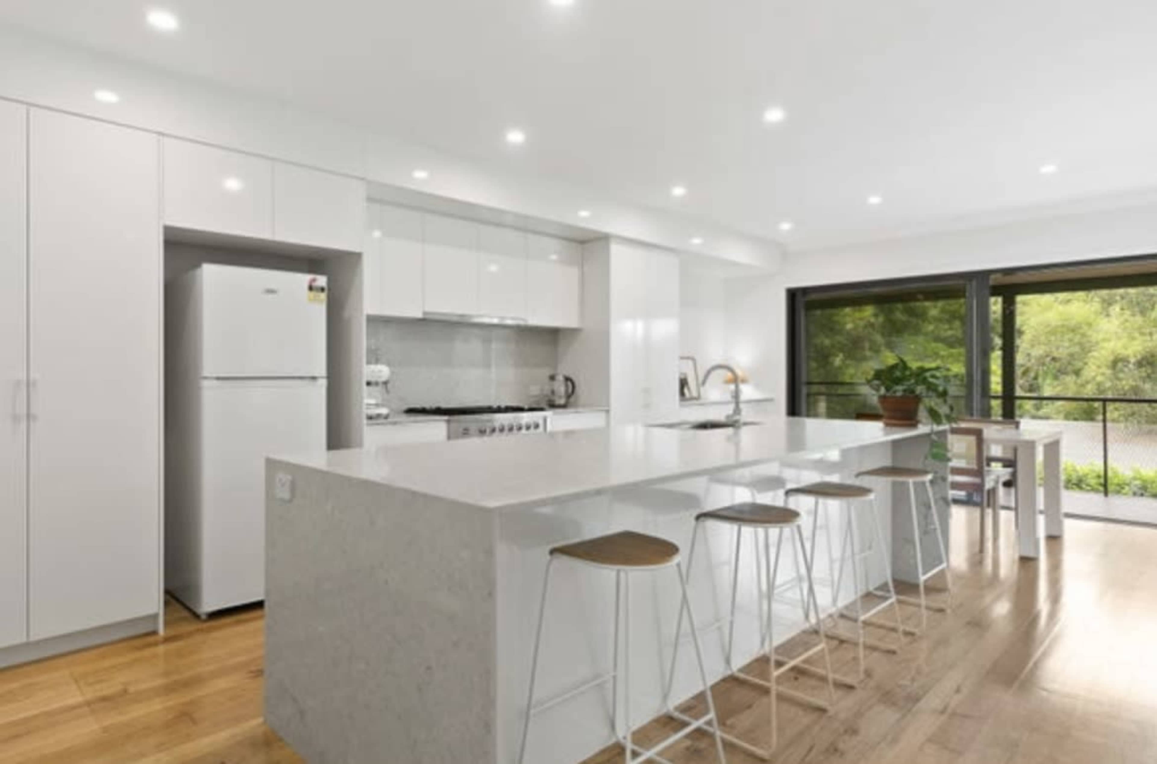 The image shows a modern kitchen with white cabinetry, a large island with bar stools, and a window overlooking a green area.