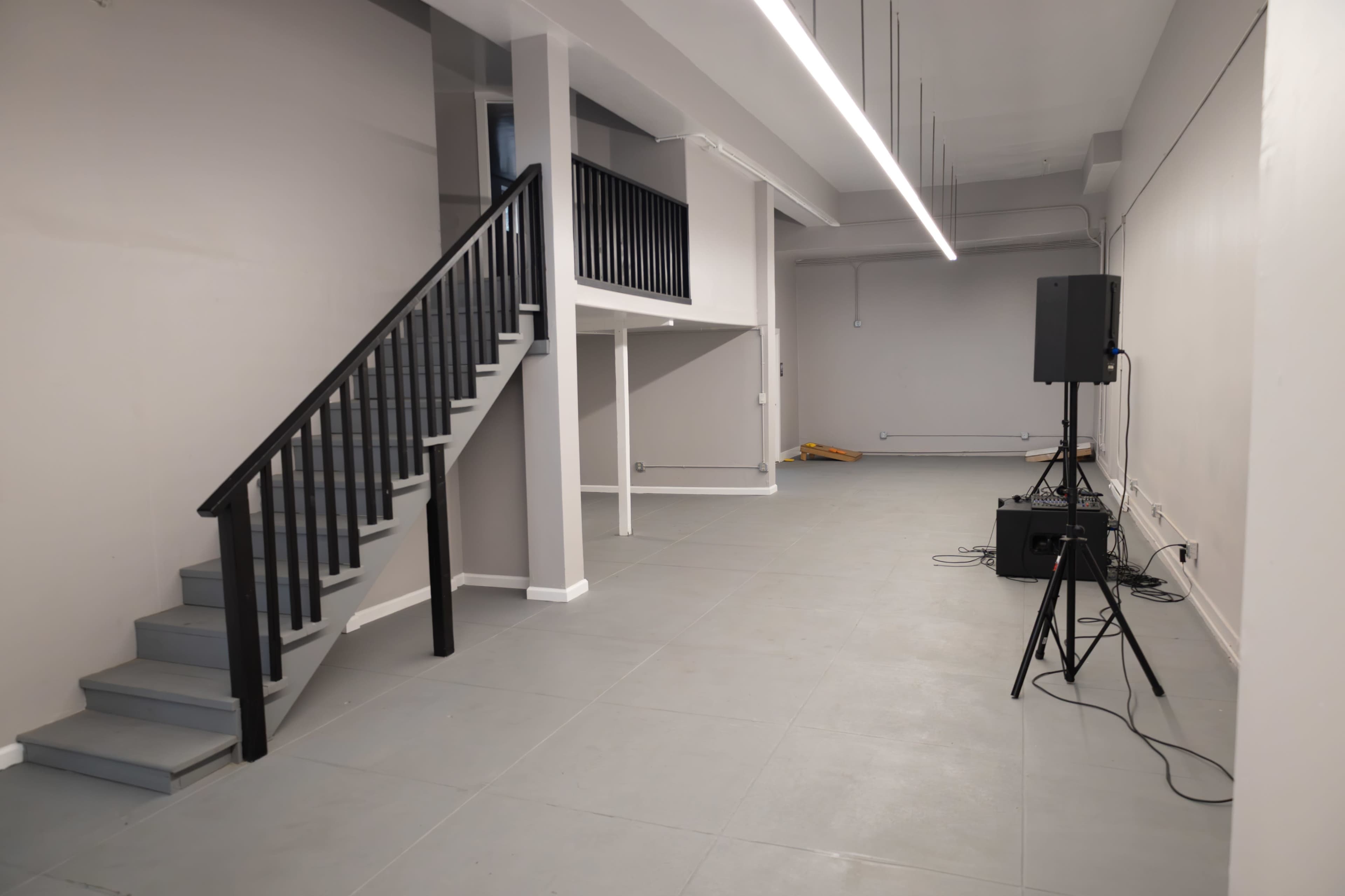 The image shows an empty, well-lit interior space with a staircase on the left and a sound system setup in the corner.