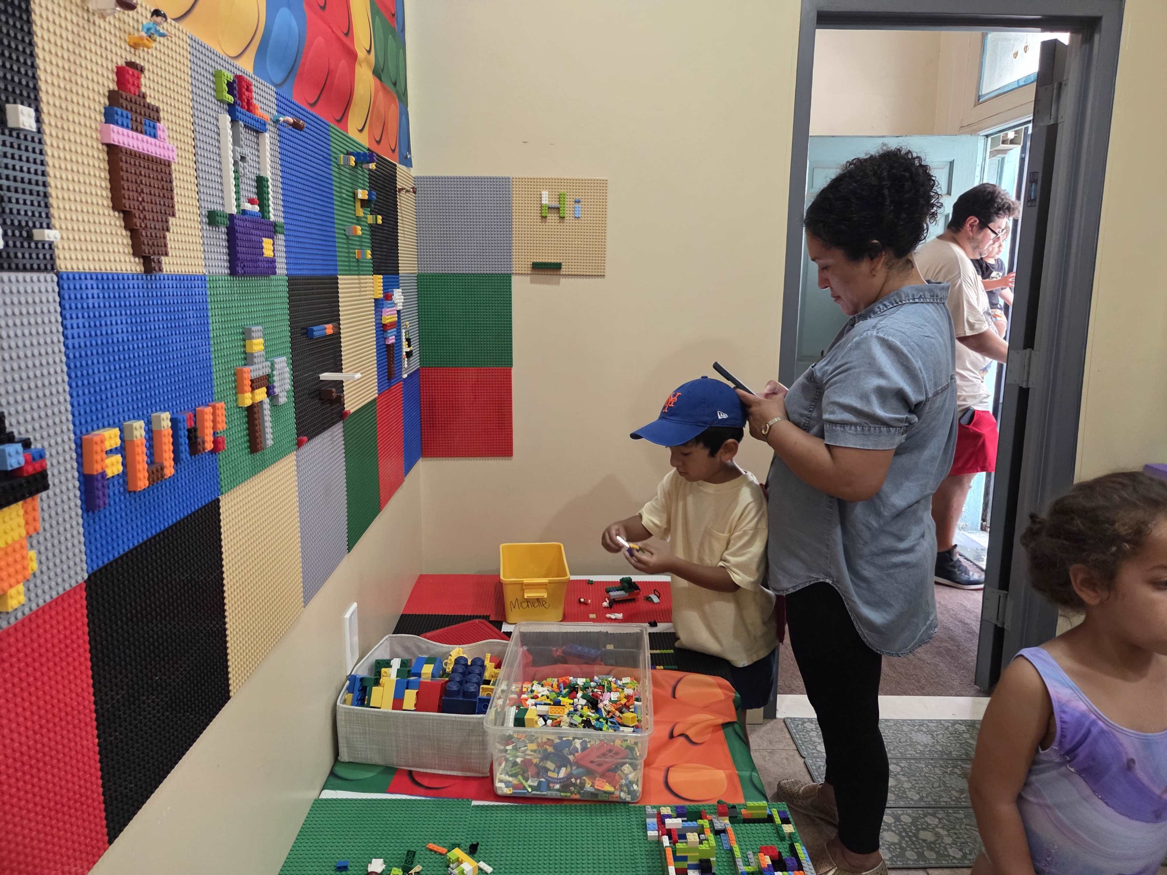 A woman stands while using her phone, watching a child play with colorful building blocks on a green mat beside a wall covered in a Lego-like display.