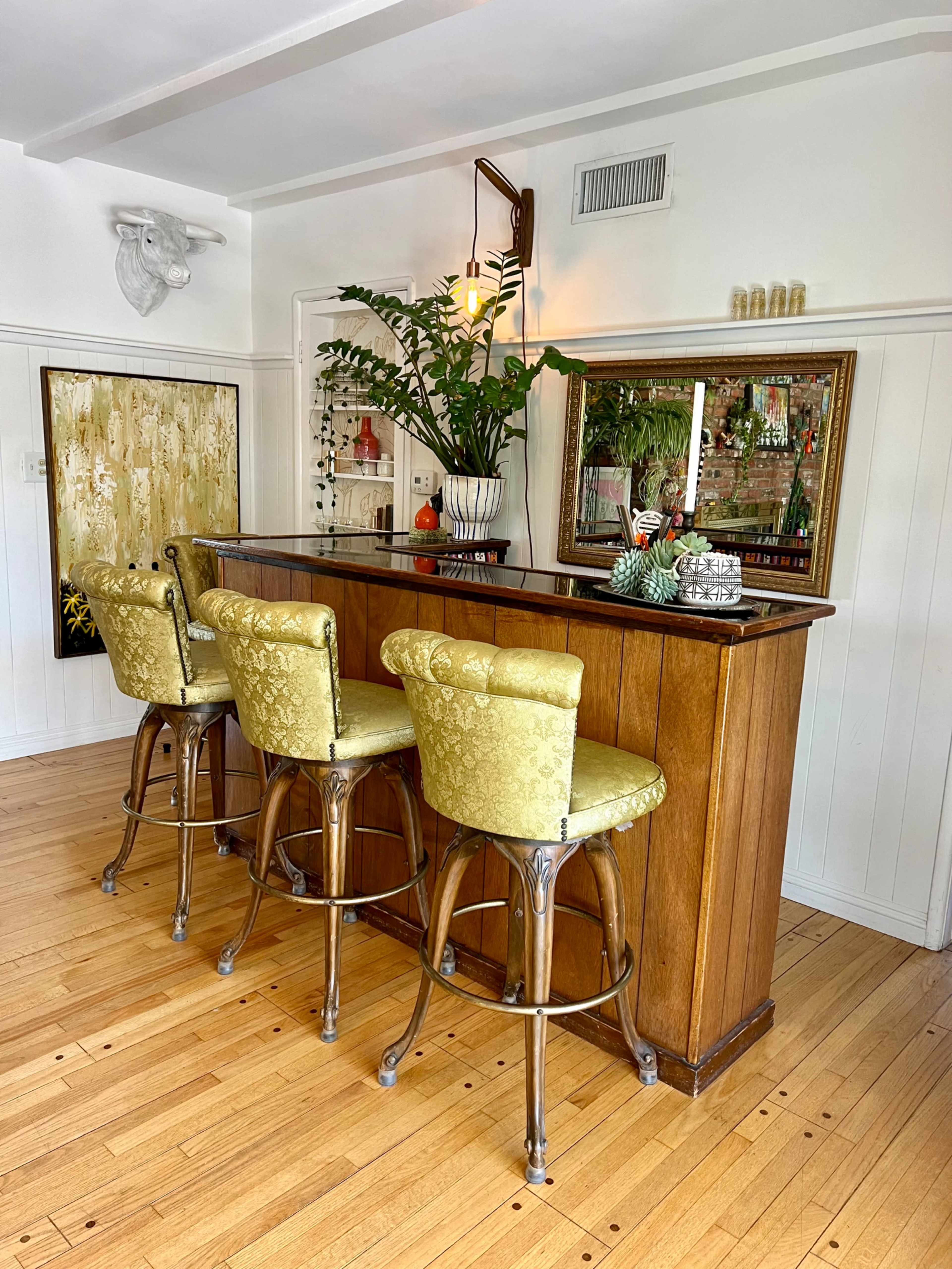 A wooden bar with three yellow upholstered stools is set against a white wall, featuring decorative plants and a mirror that reflects various bottles on the back shelf.