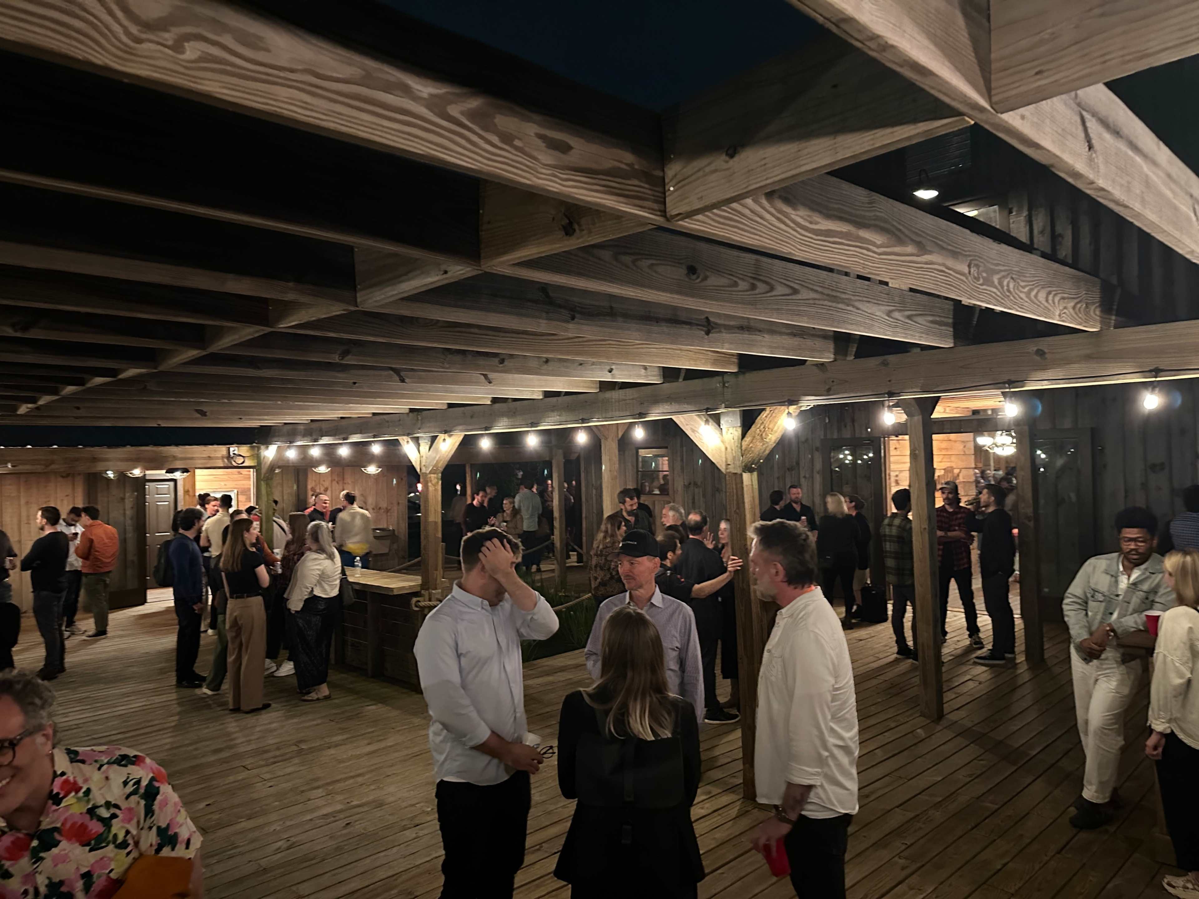 A large gathering of people socializes under a wooden pavilion with string lights at night.