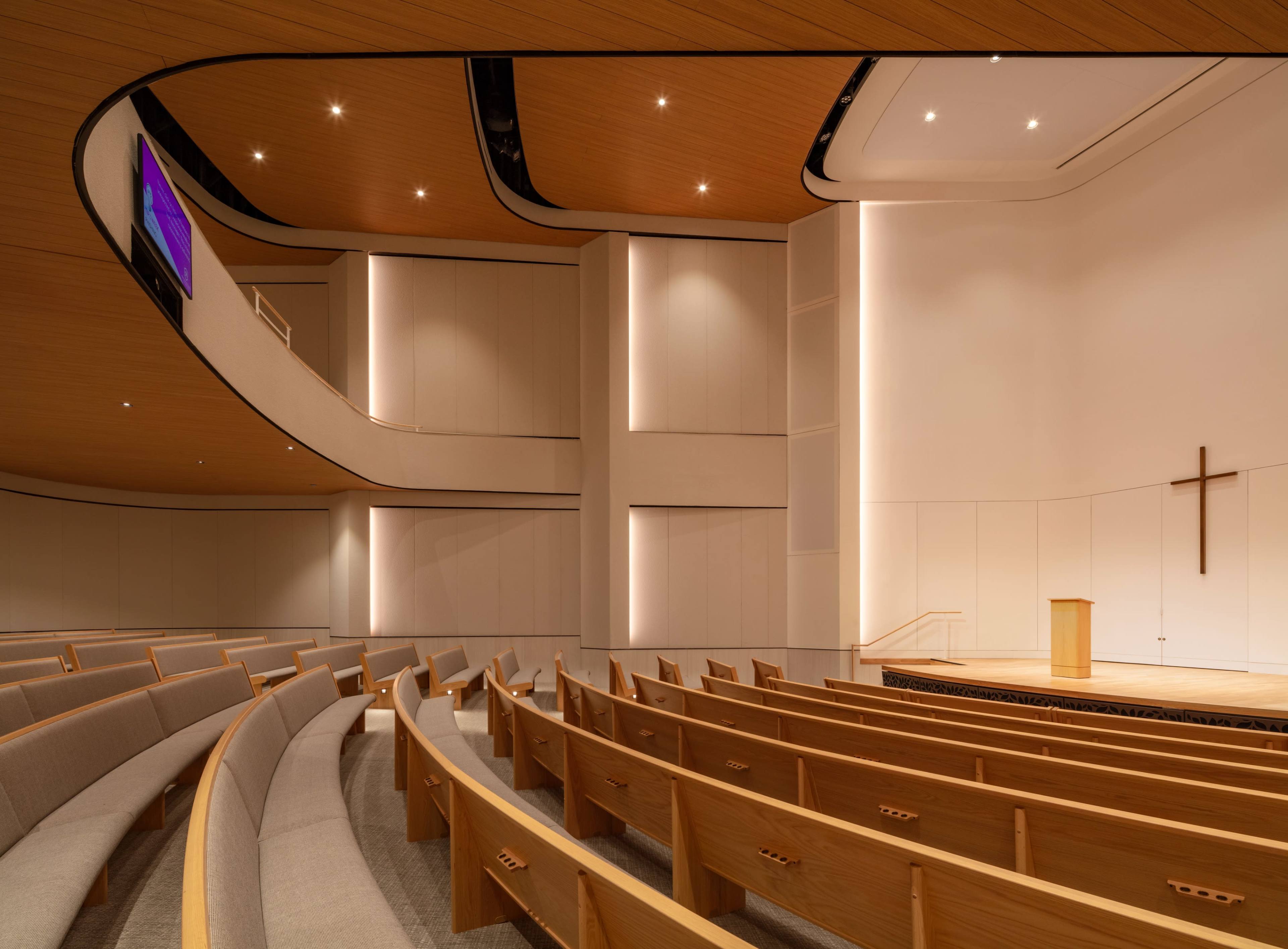 The image shows a modern interior of a church with curved seating, a wooden podium, and a large cross on the wall.