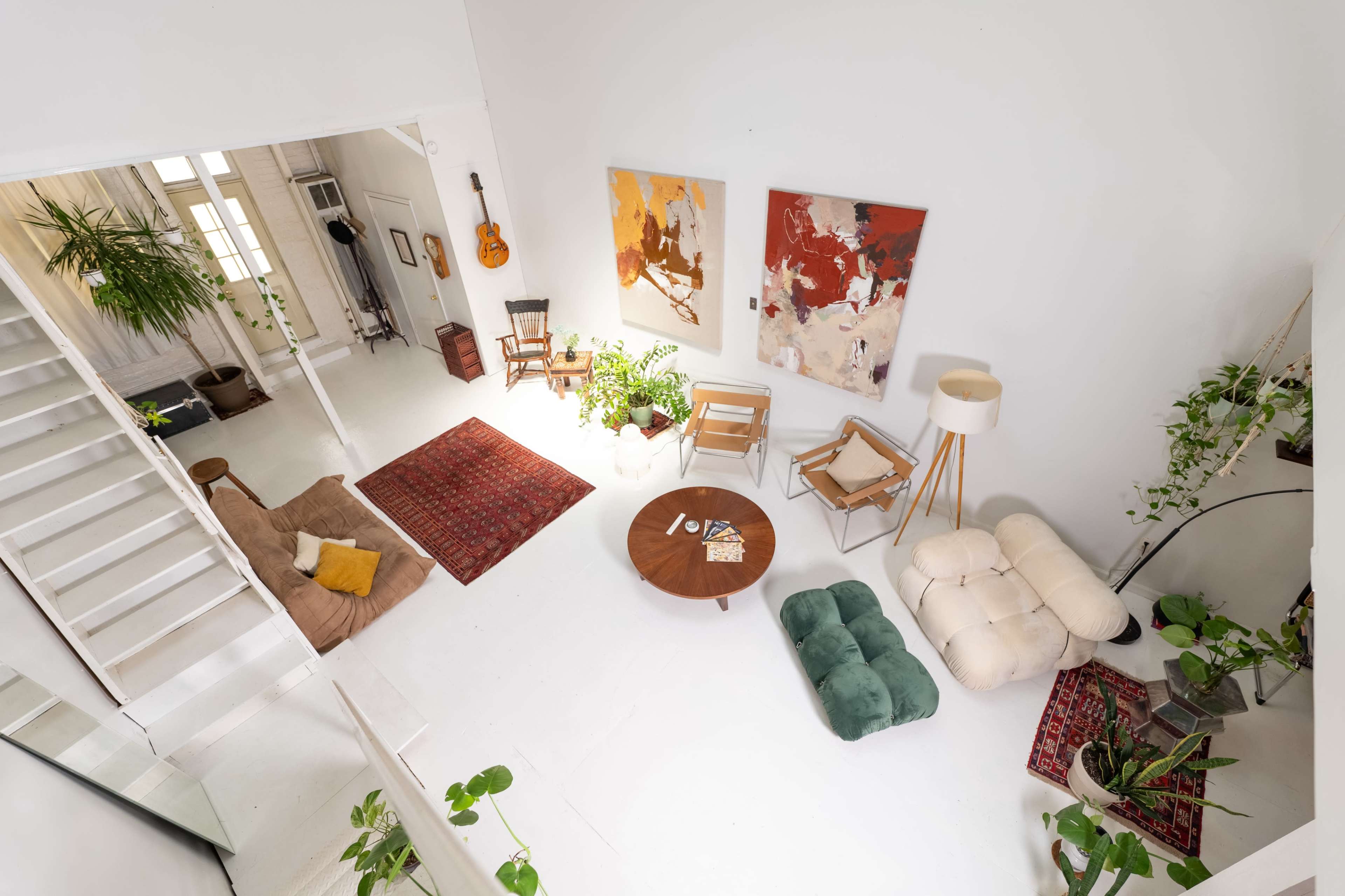 The image shows a bright, open living space featuring a mix of furniture, plants, and artwork on the walls, with a staircase visible in the background.