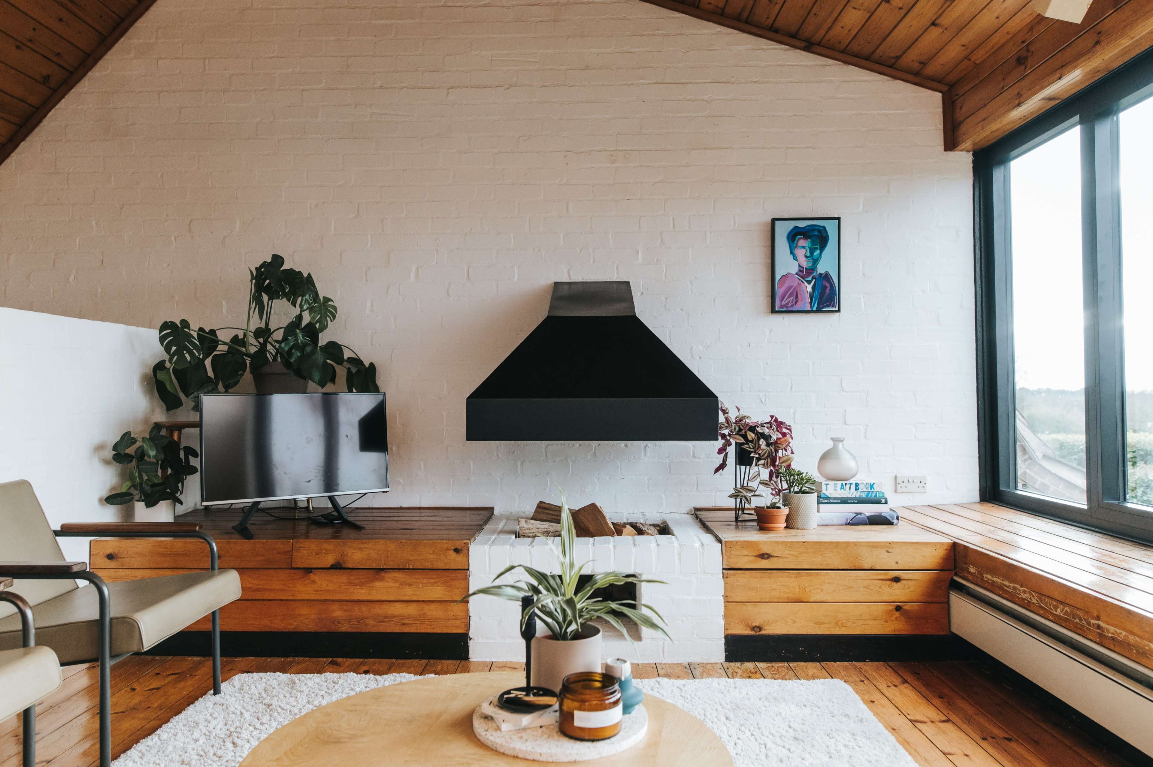 A modern living room features a flat-screen TV, a black chimney, a round coffee table, and large windows with a view, surrounded by plants and wooden accents.