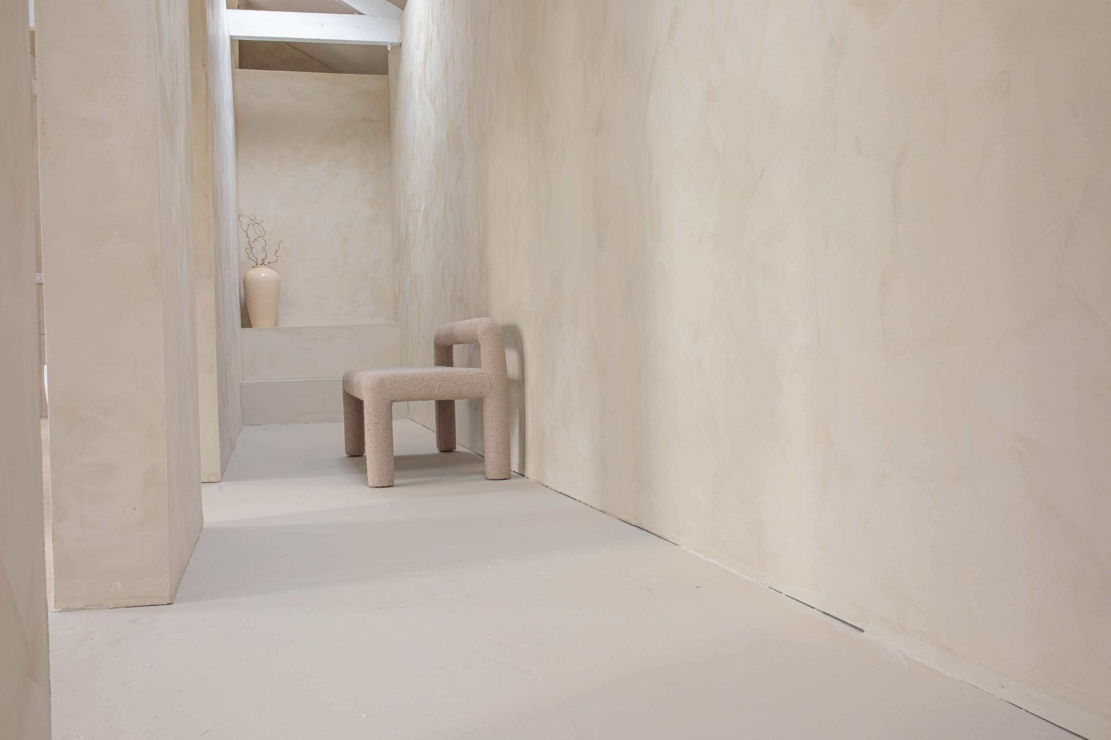 A narrow, softly lit hallway features a minimalist chair and a vase on a ledge against textured, light-colored walls.