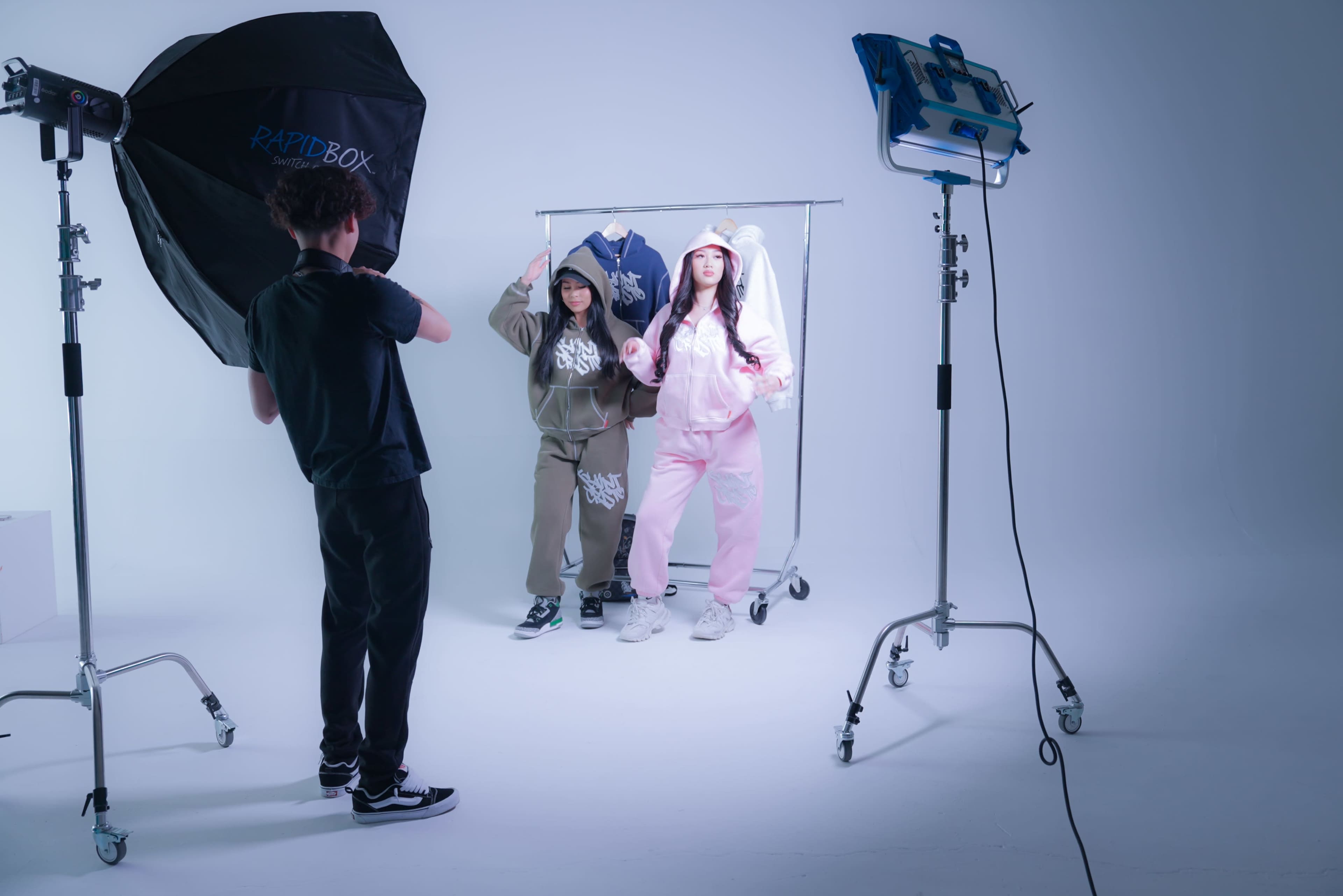 Two models, one in olive green and the other in pink, pose in front of a clothing rack in a photo studio while a photographer sets up lighting equipment.