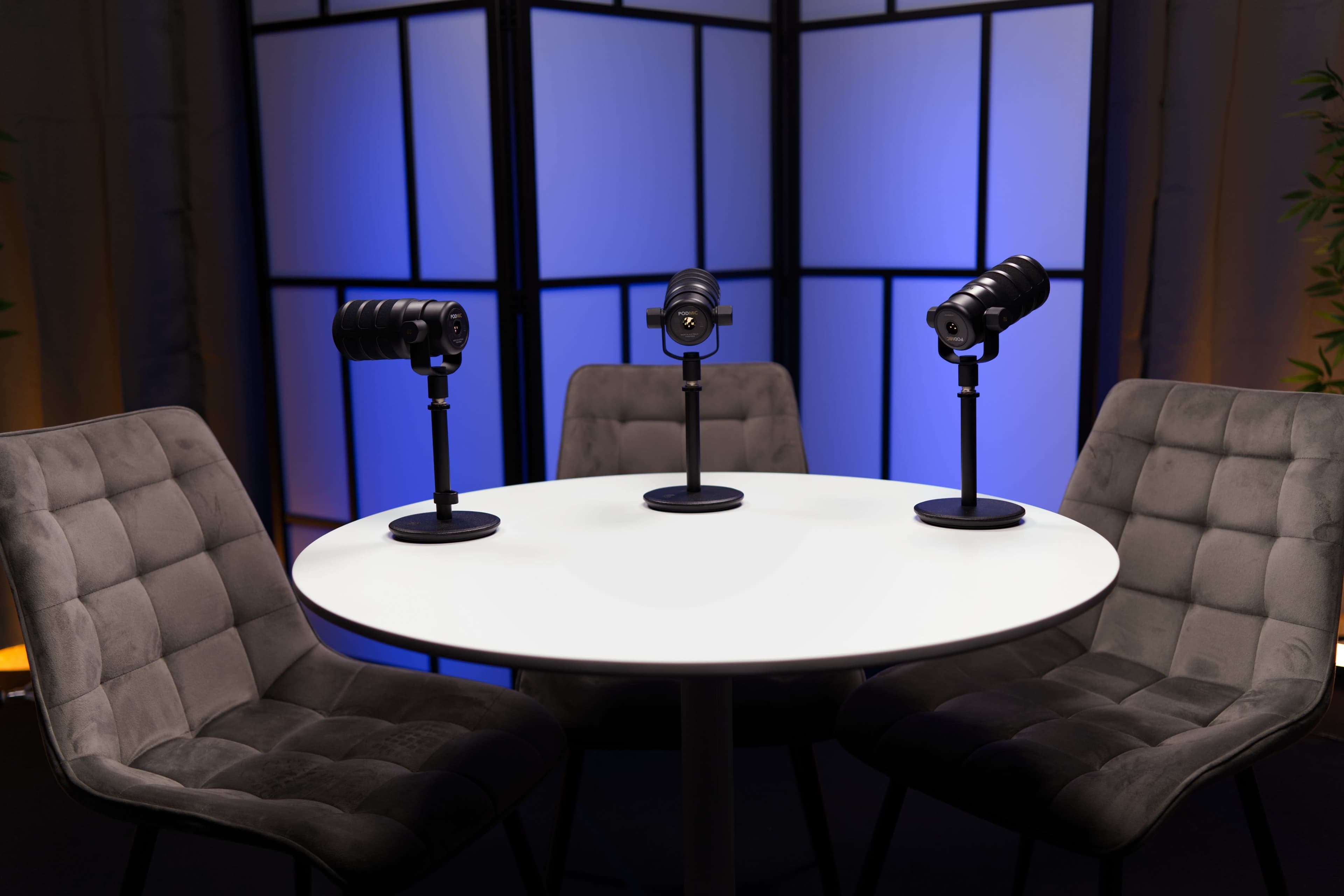 Three cameras on stands are positioned in front of a round white table, with a decorative backdrop featuring blue and black screens behind them.