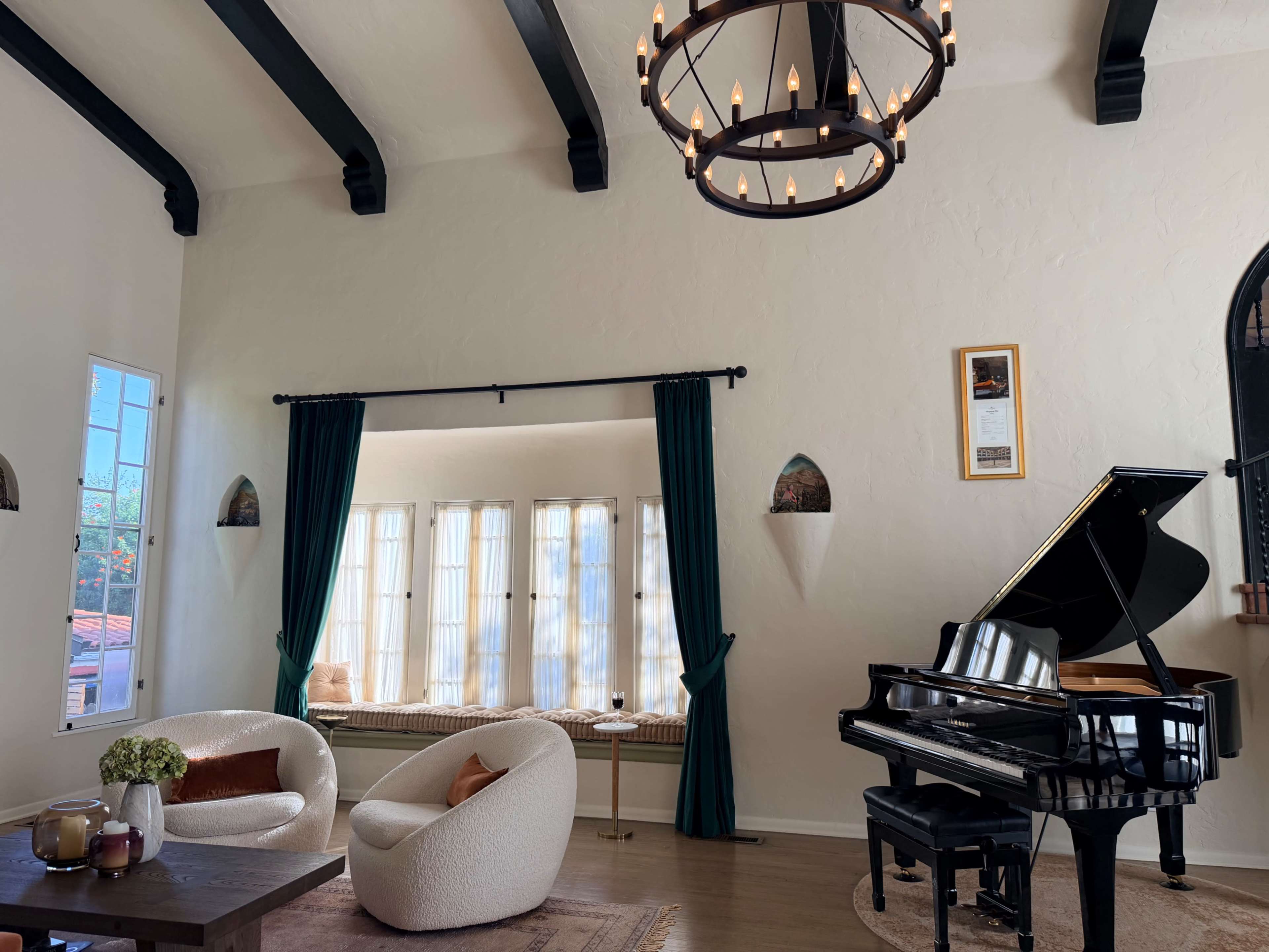 The image shows a cozy interior space featuring a grand piano, two upholstered chairs, and large windows adorned with green curtains.
